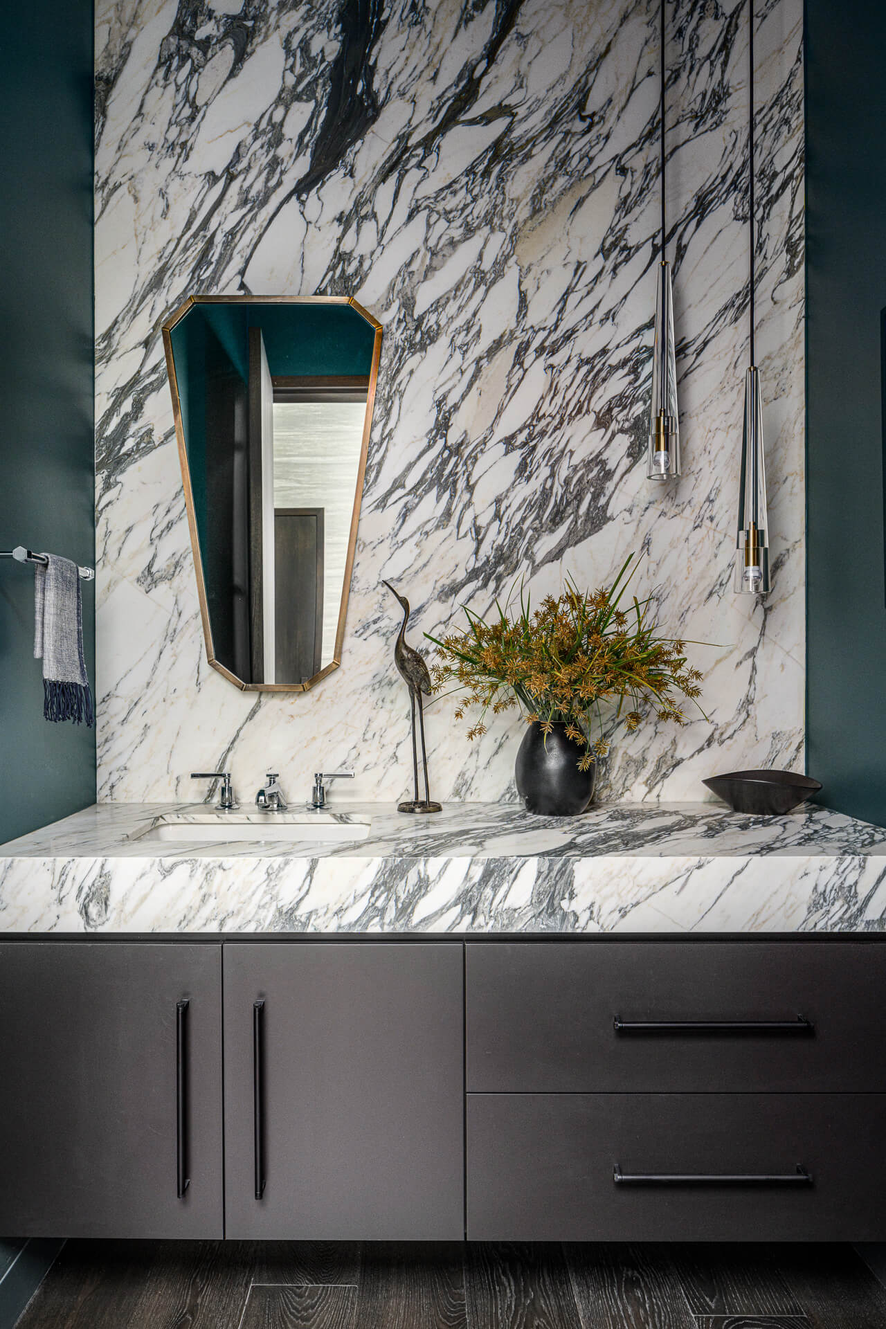 Marble bathroom vanity with dark cabinetry, geometric mirror, and a vase of flowers.