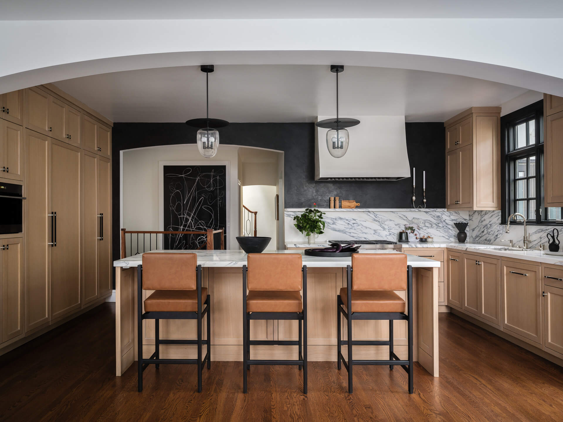 Kitchen with natural wood cabinetry, marble island, three leather stools, and black-framed windows.