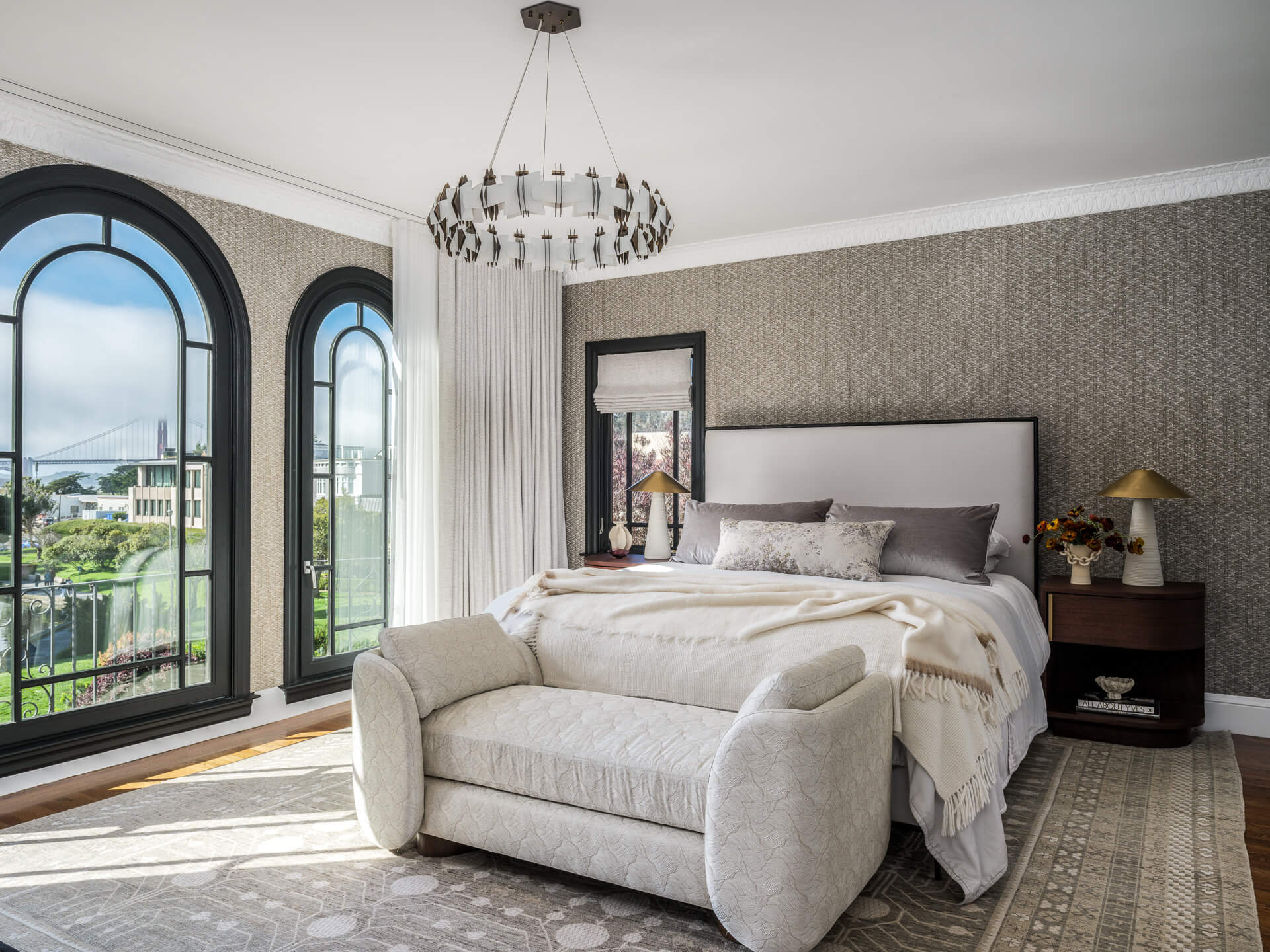 Serene bedroom with upholstered bed, curved bench, arched windows, and crystal chandelier.