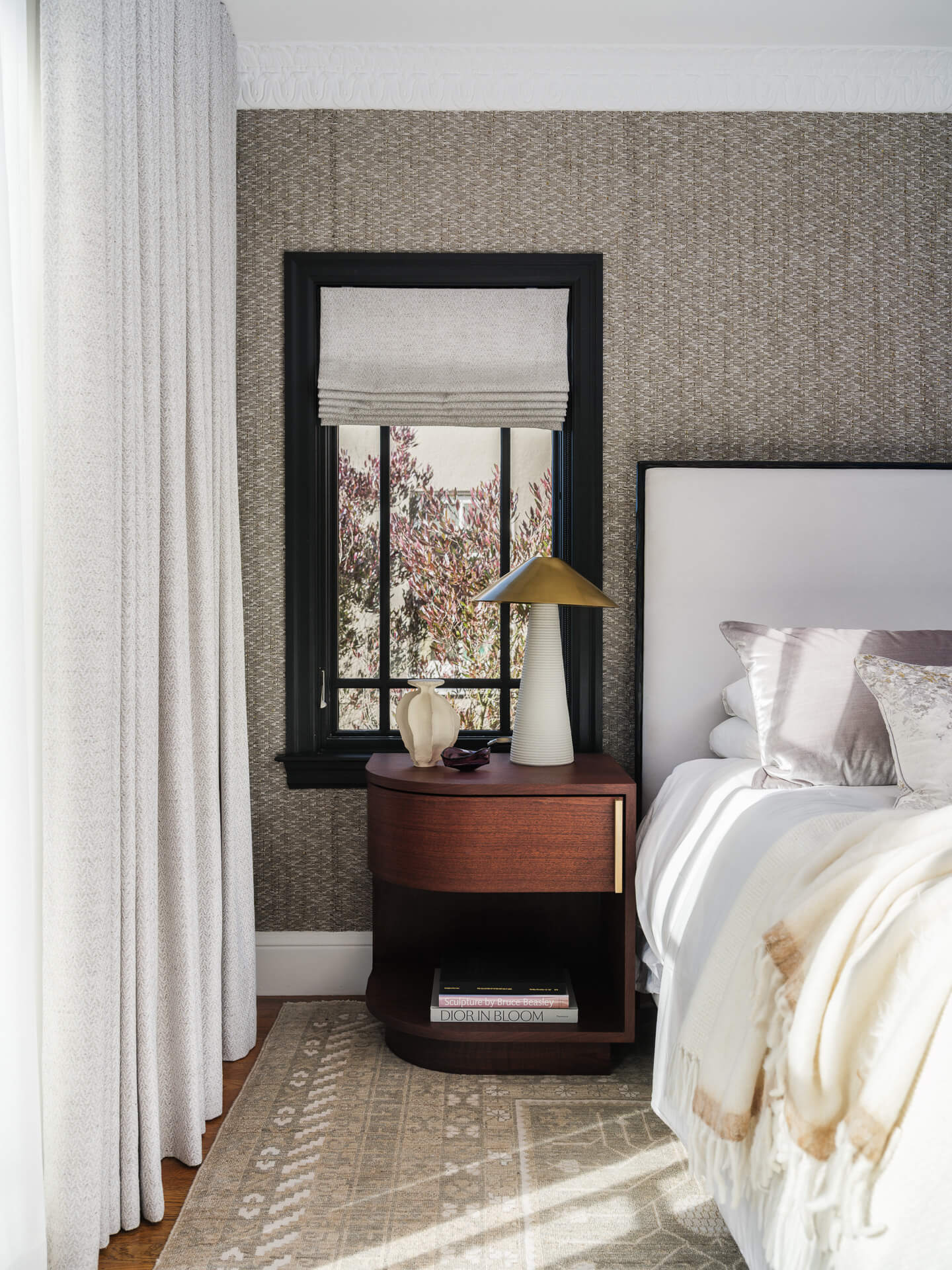 Bedroom detail with upholstered bed, wood nightstand, brass lamp, and tall black-trimmed window.
