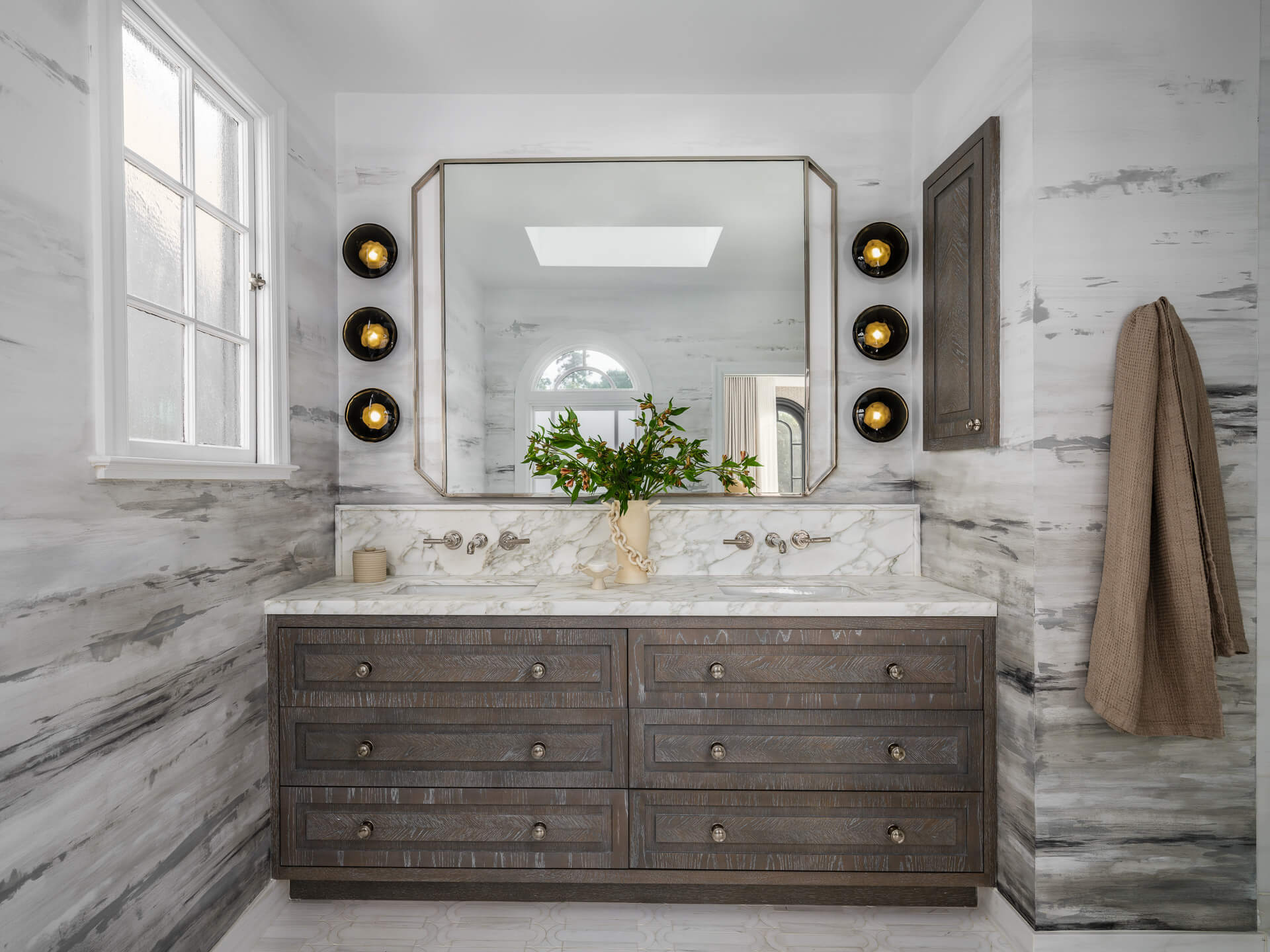 Marble bathroom with wide wood vanity, large framed mirror, black-and-gold sconces, and flowers.