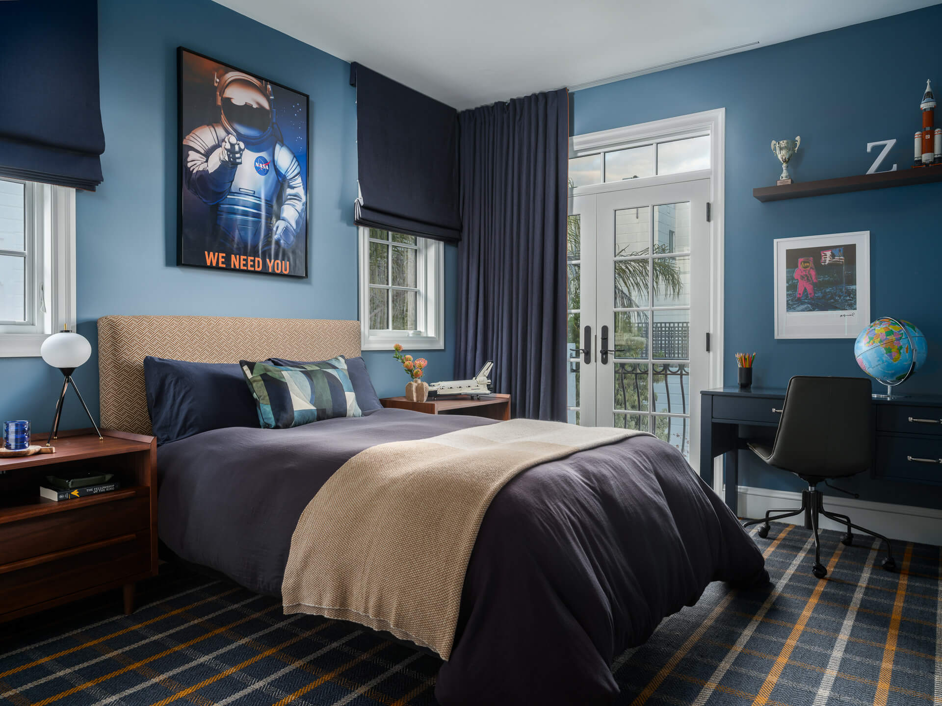 Blue bedroom with upholstered bed, plaid rug, desk, dark curtains, and framed astronaut artwork.