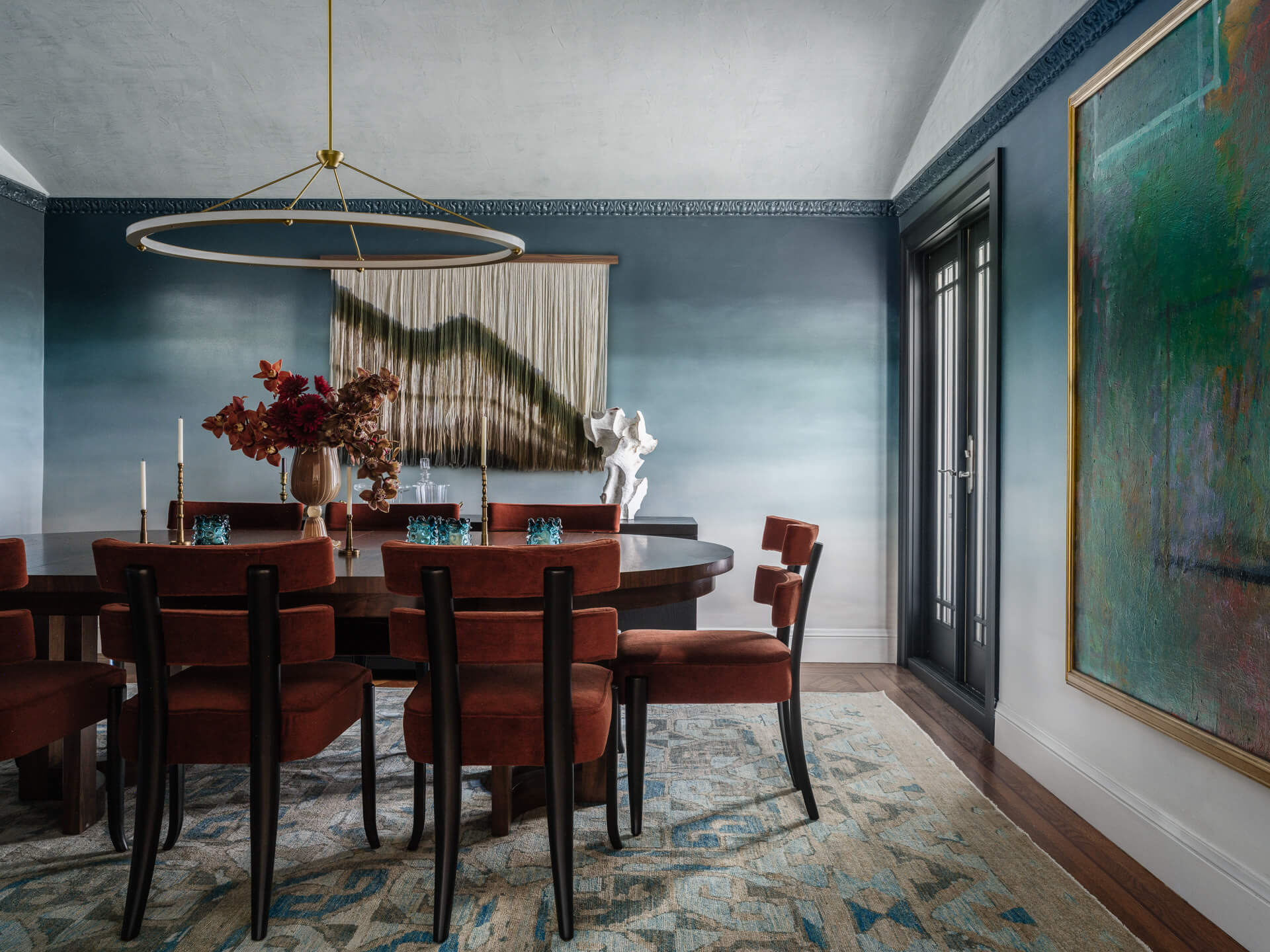 Moody dining room with blue-gray walls, rust-colored chairs, oversized abstract artwork, and sculptural pendant light.