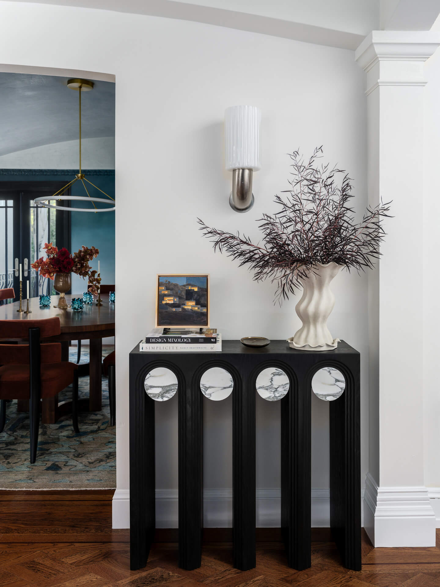 Sculptural black console topped with a white vase of branches beneath a ribbed wall sconce.