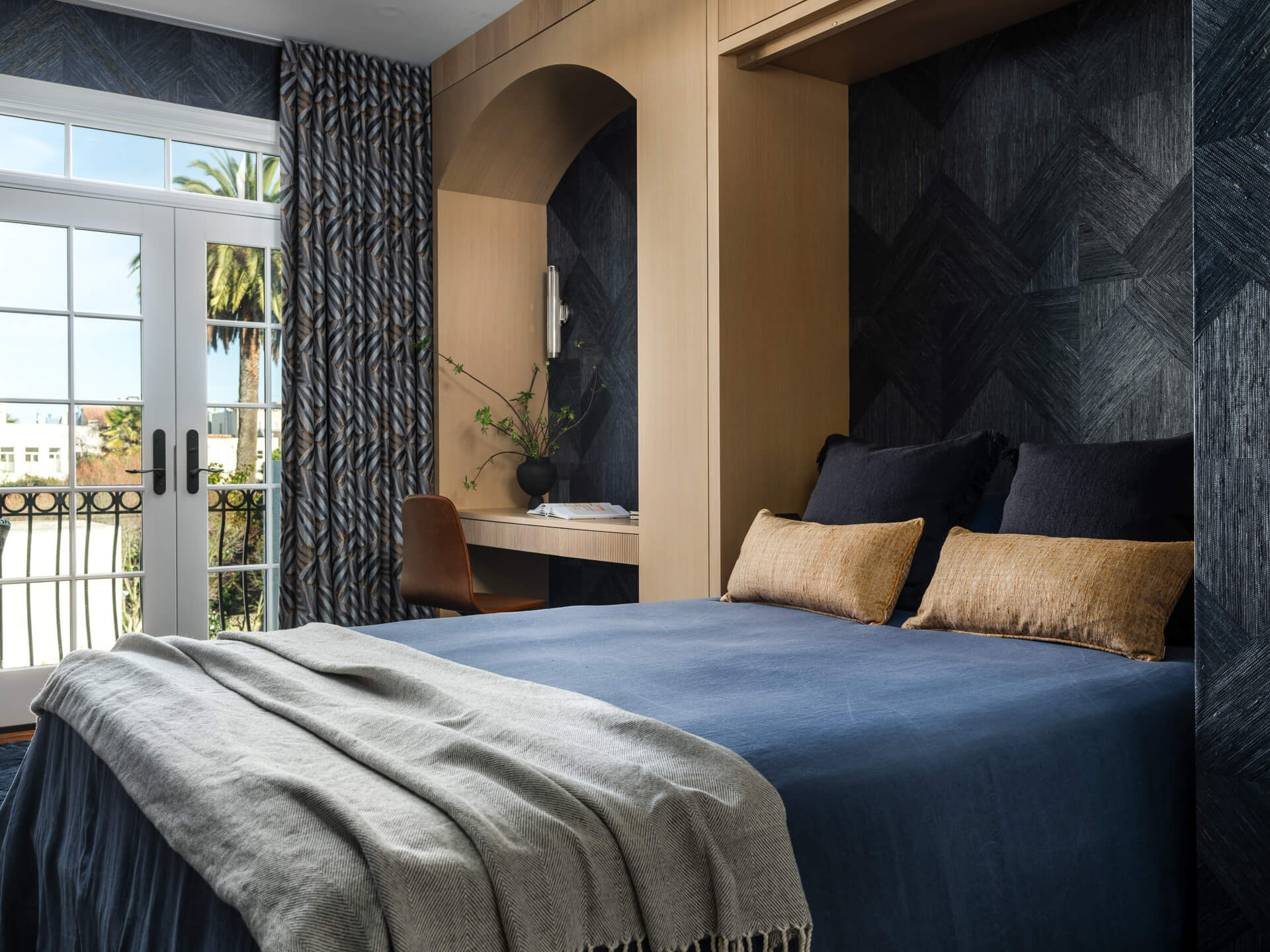 Dark blue bedroom with wood-framed alcove, built-in desk, navy bedding, and French doors opening to a balcony.