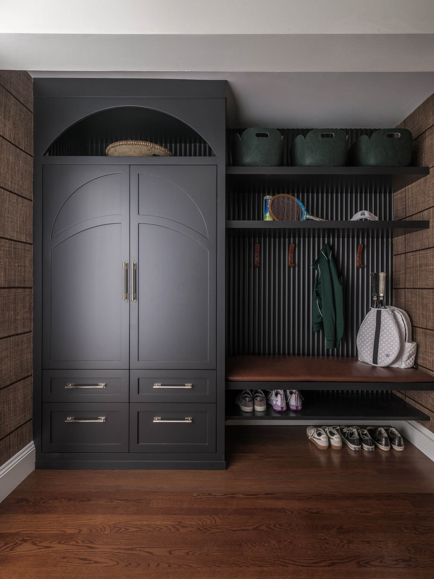 Dark built-in mudroom with closed cabinetry, open shelves, bench, hanging storage, and shoe racks.