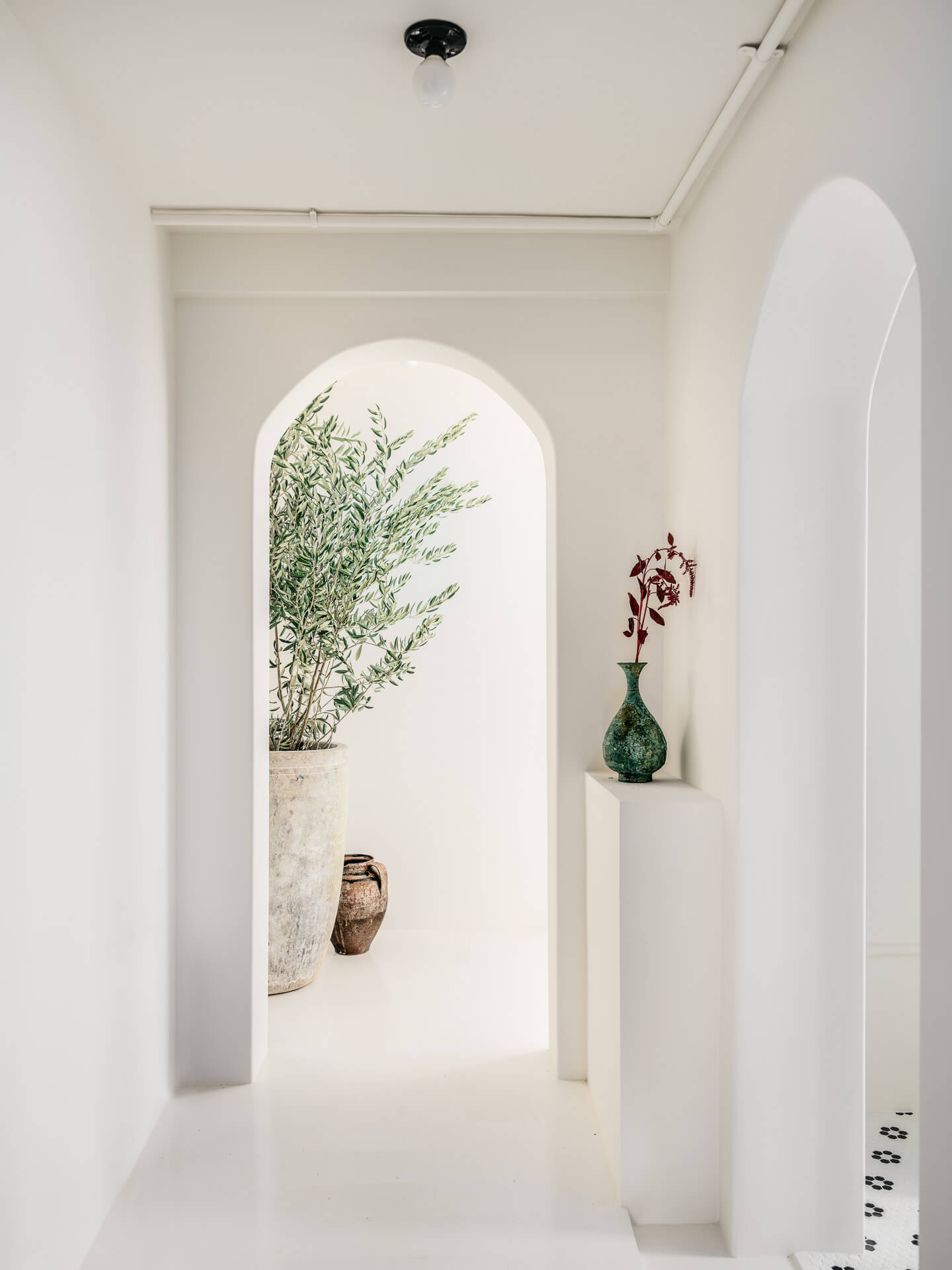 Serene white hallway with rounded archways, small display niches, and potted greenery beyond.