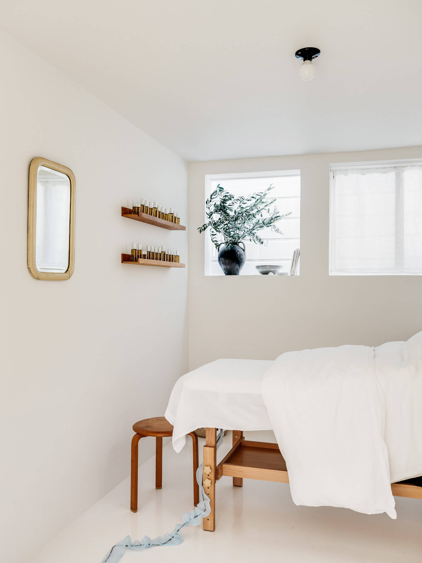 Minimal treatment room with white-covered massage table, small wood stools, wall mirror, and shelves of bottles.