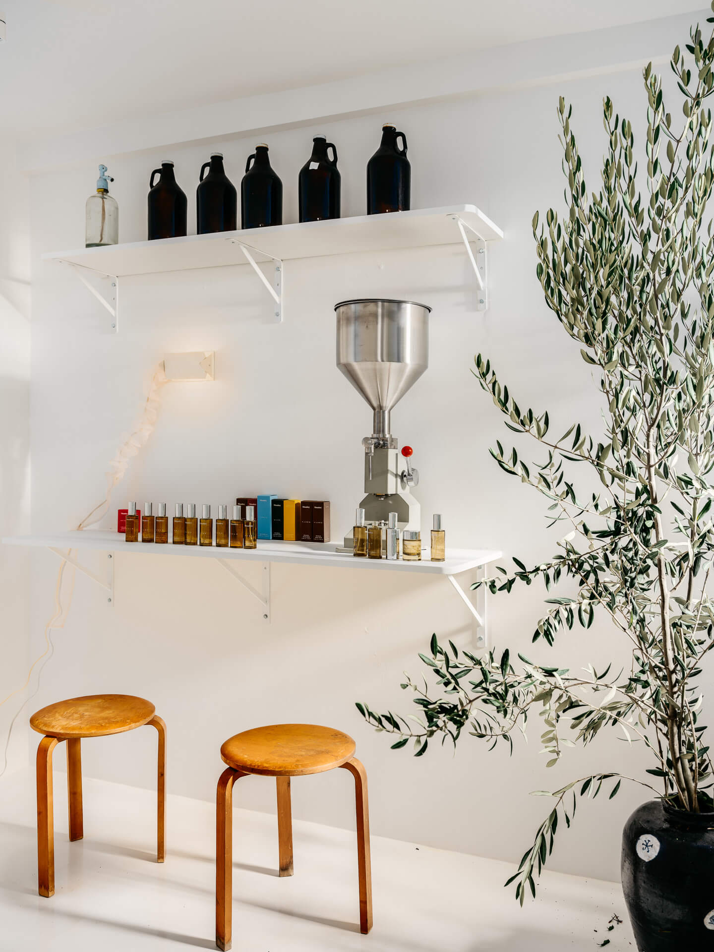 White beverage bar with glass bottles, countertop dispenser, wood stools, and a large potted olive tree.