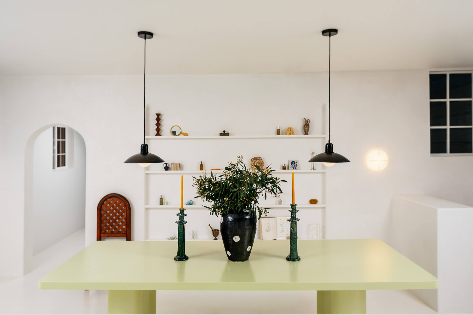 Pale green communal table with black vase and candlesticks beneath pendant lights in a bright white room.