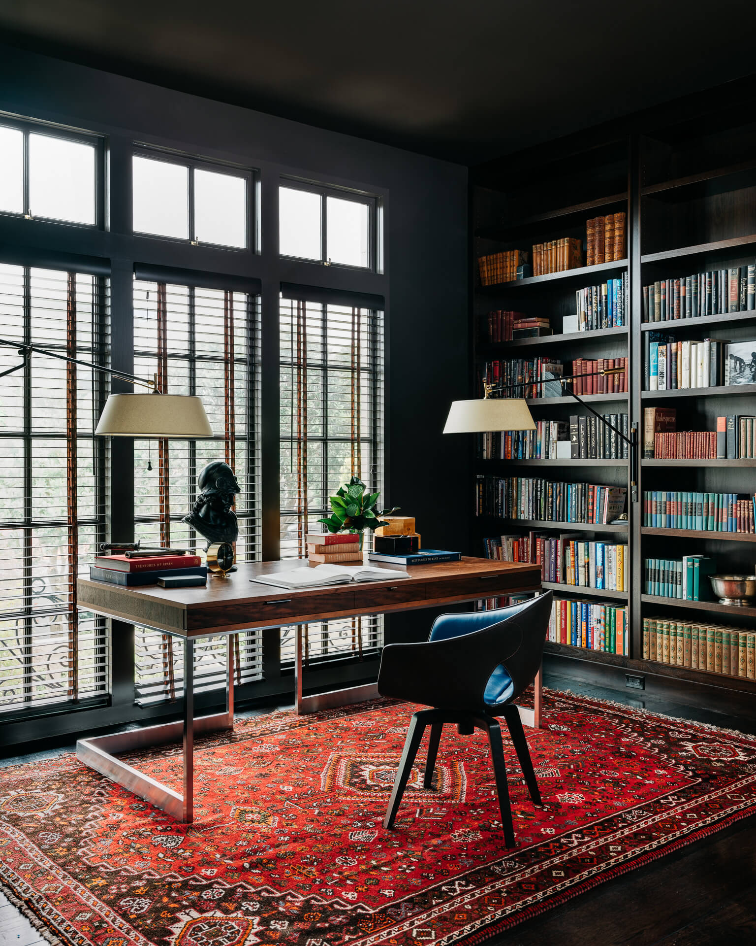 Dark home office with wall-to-wall bookshelves, large windows, wood desk, and a red patterned rug.