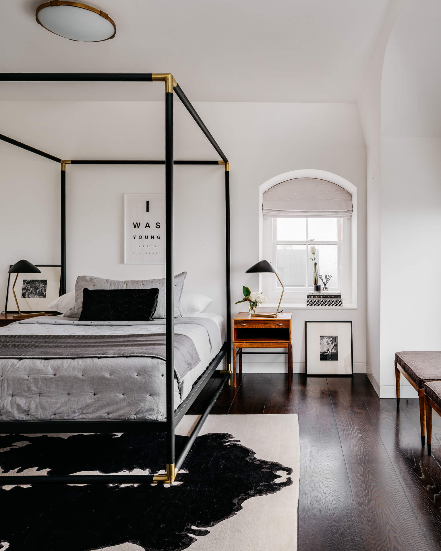Bright bedroom with black metal canopy bed, arched window, dark wood floors, and a black hide rug.