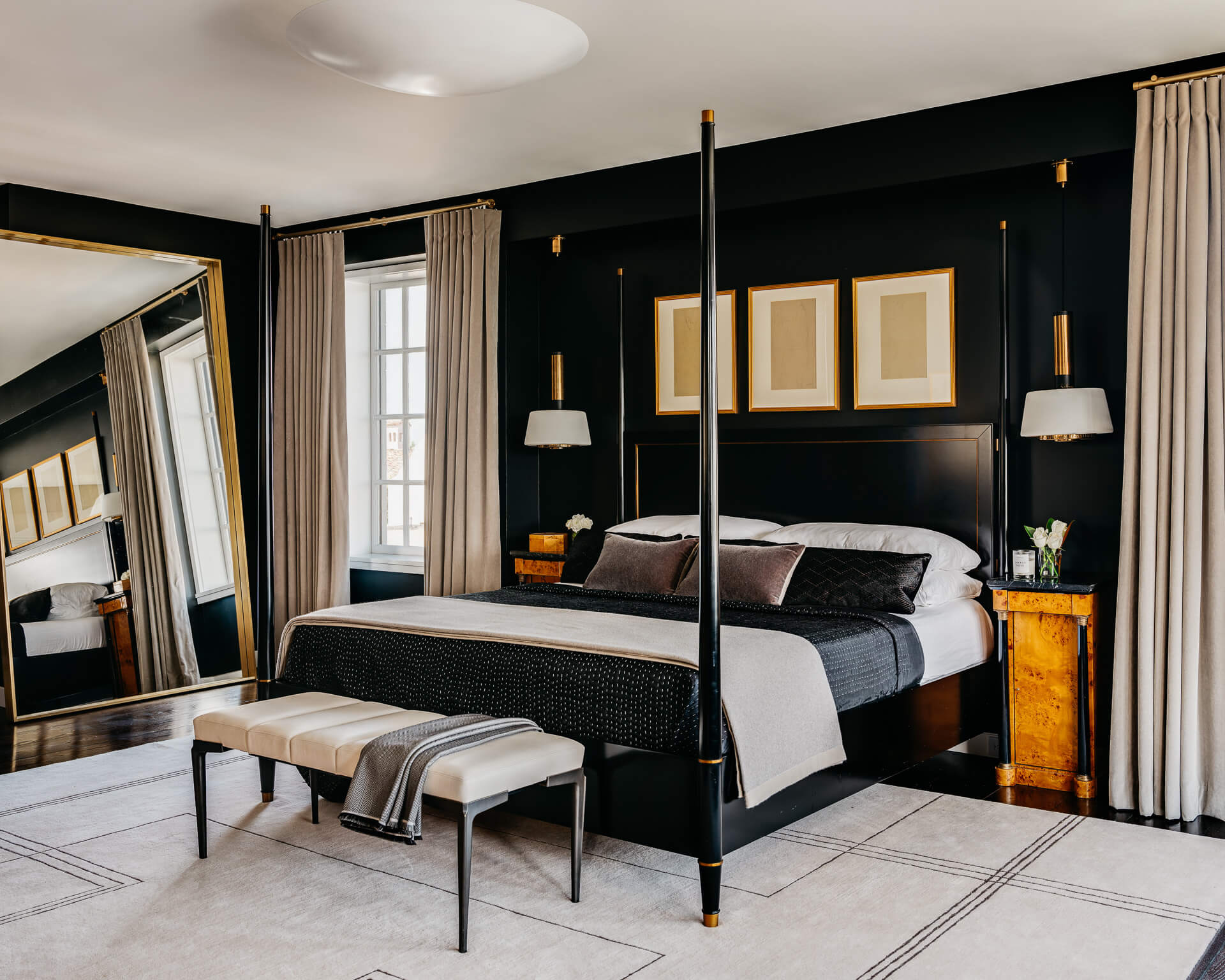 Dark bedroom with black canopy bed, large leaning mirror, wood nightstands, and tall curtained windows.
