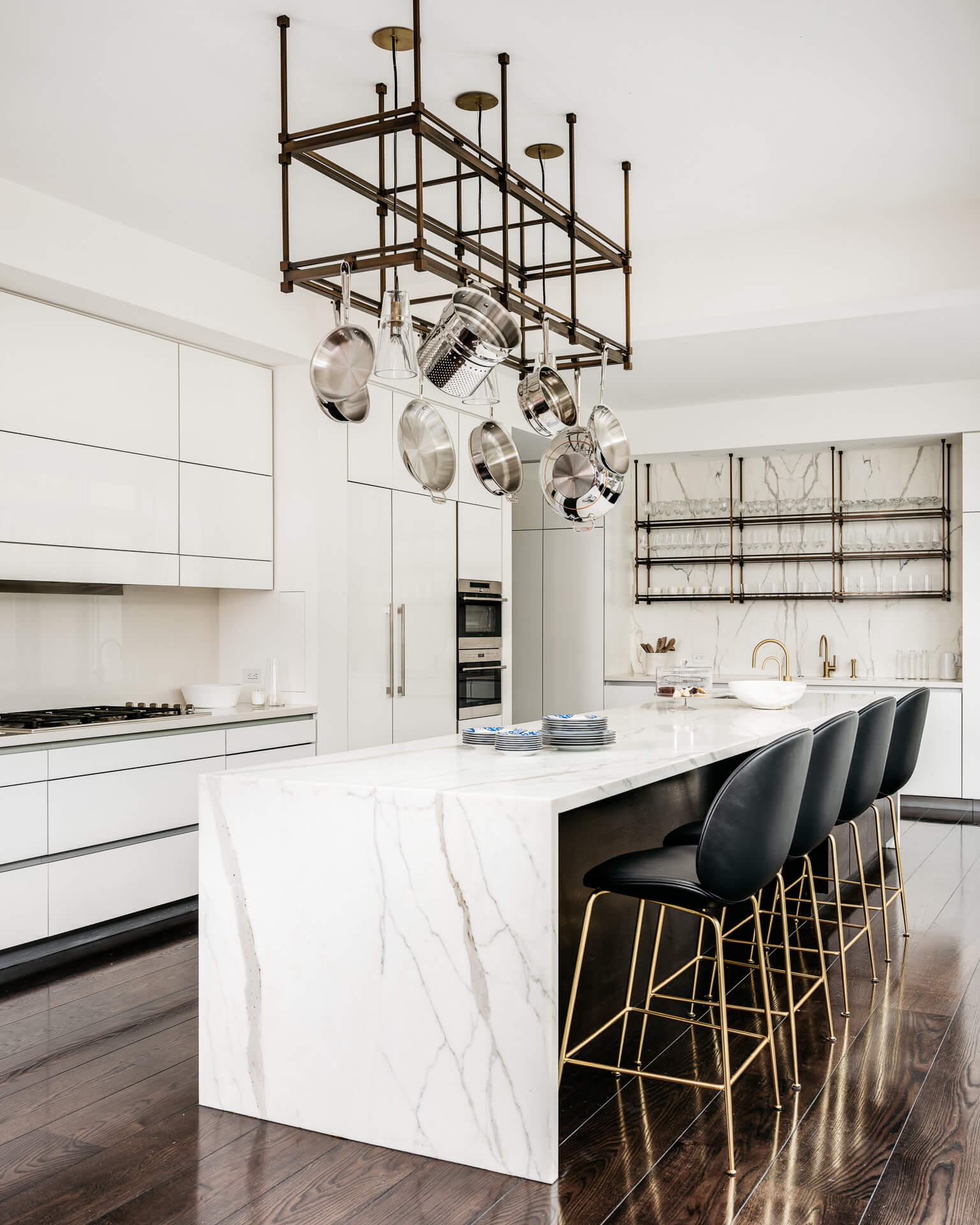 White kitchen with marble waterfall island, black bar stools, and an overhead hanging rack.