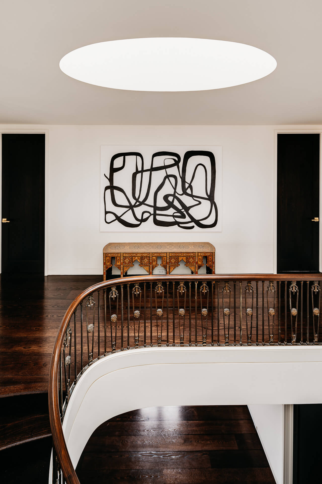 Curved stair landing with dark wood floors, oval skylight, and abstract artwork above a console.