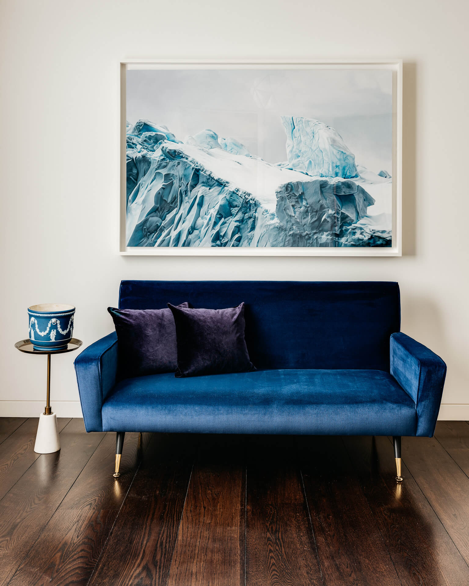 Deep blue velvet sofa beneath a large iceberg painting against a pale wall.