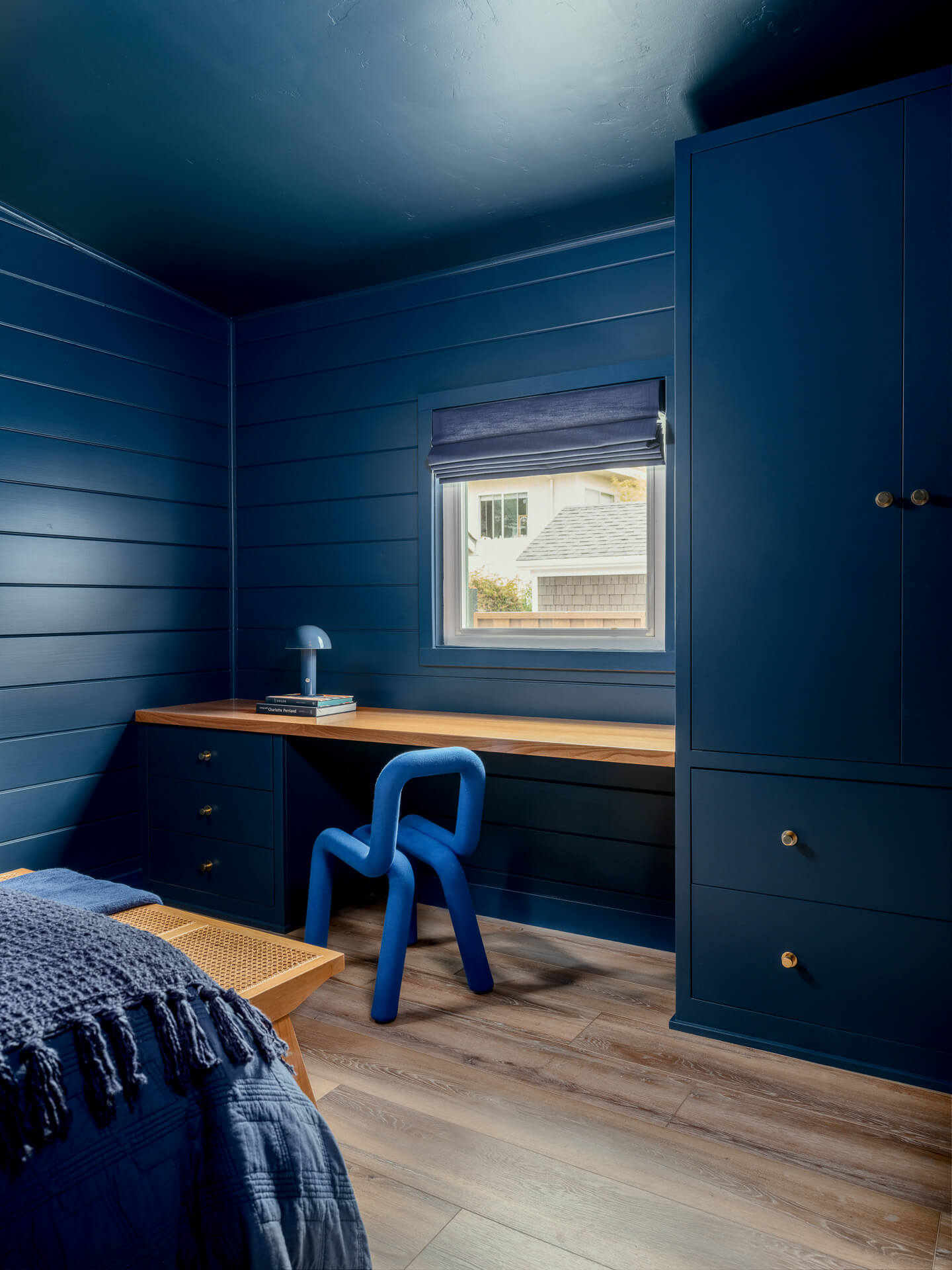 Deep blue bedroom corner with built-in wardrobe, wood desk, bright blue chair, and small window.