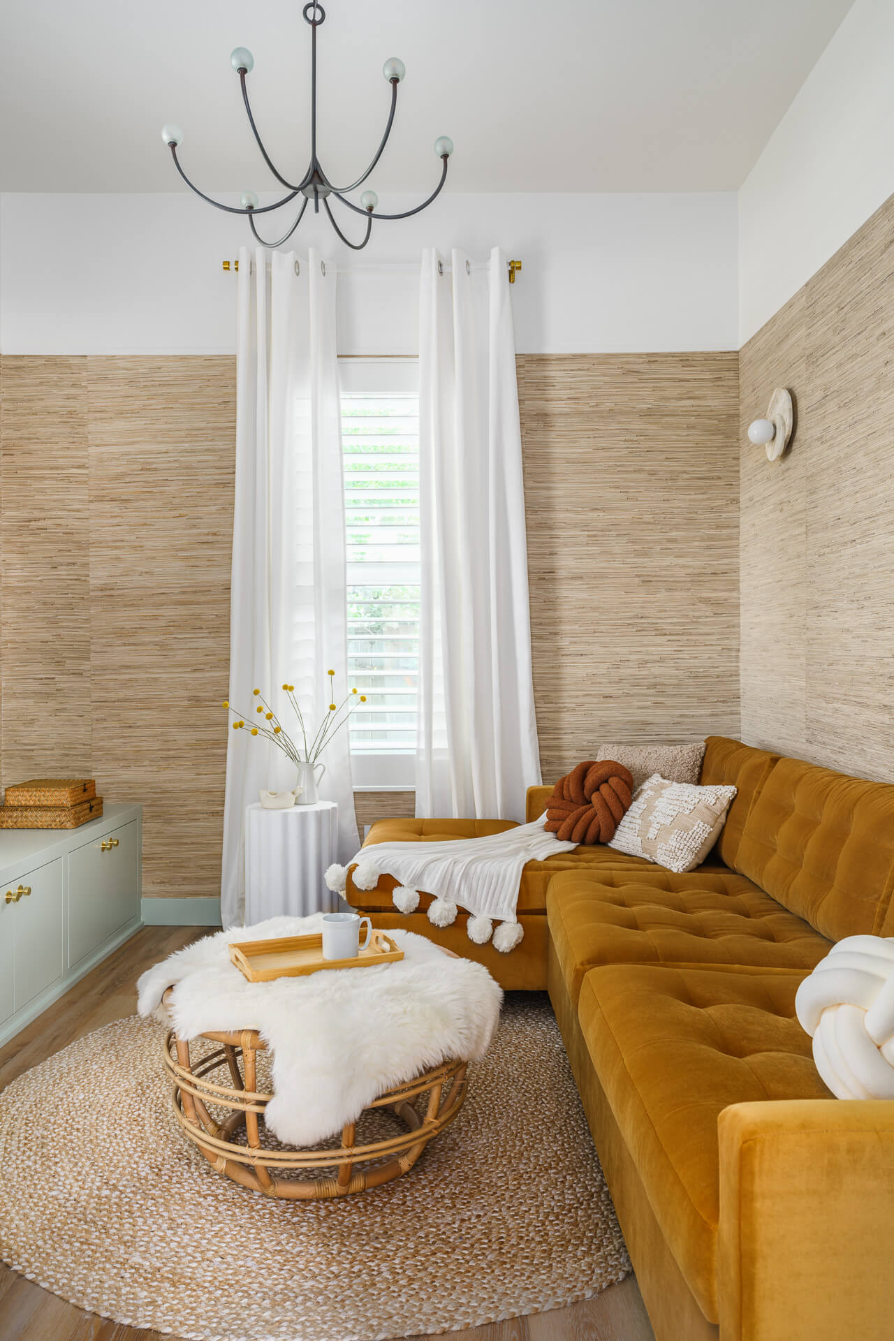Bright sitting room with mustard sectional, woven coffee table, textured walls, and tall curtained window.