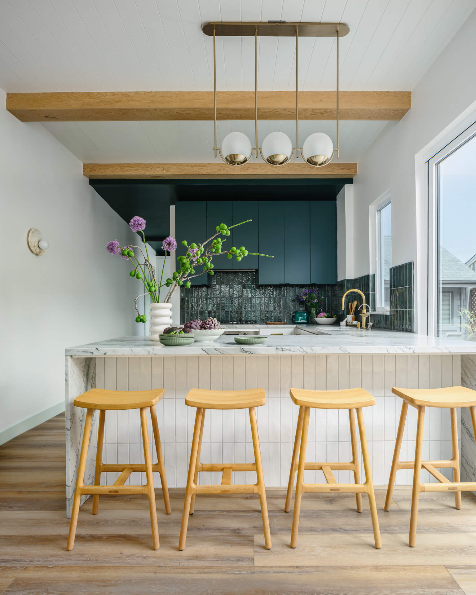 White kitchen island with four wood stools, deep green cabinetry, globe pendant lights, and exposed ceiling beams.