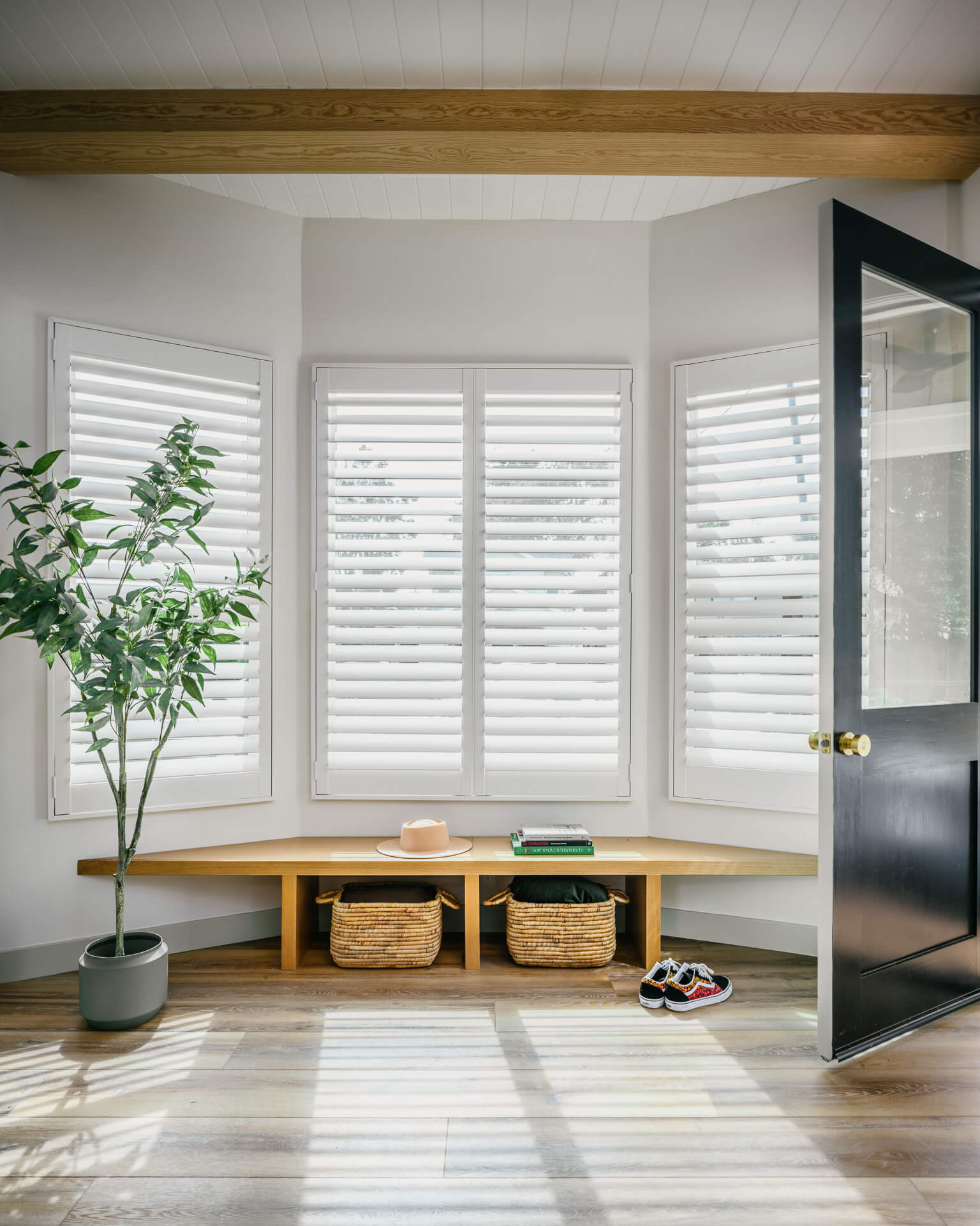 Sunlit entry nook with wraparound windows, built-in wood bench, potted tree, and black door.