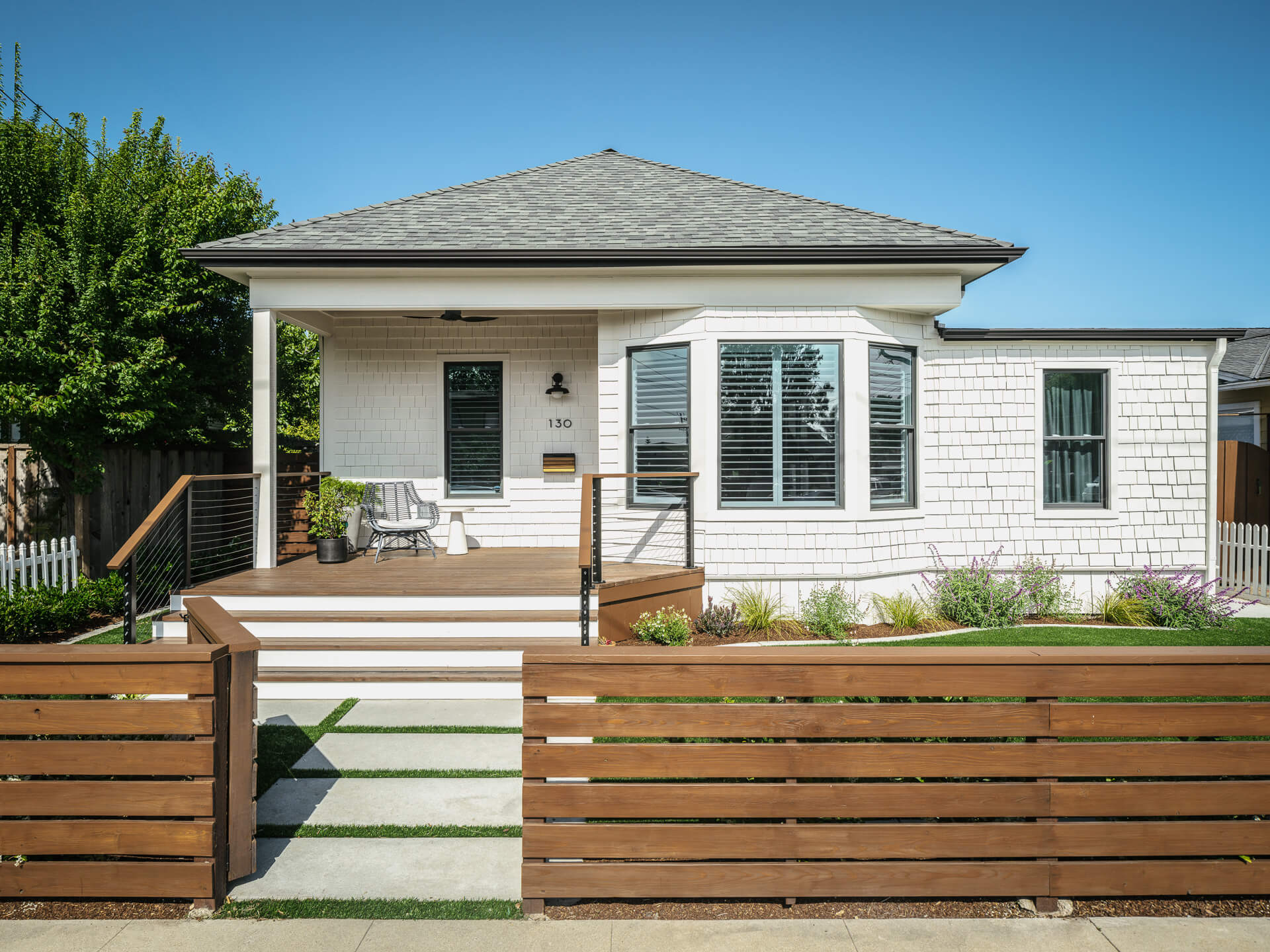 White shingle beach house with front porch, wood fence, landscaped yard, and stepping-stone walkway.