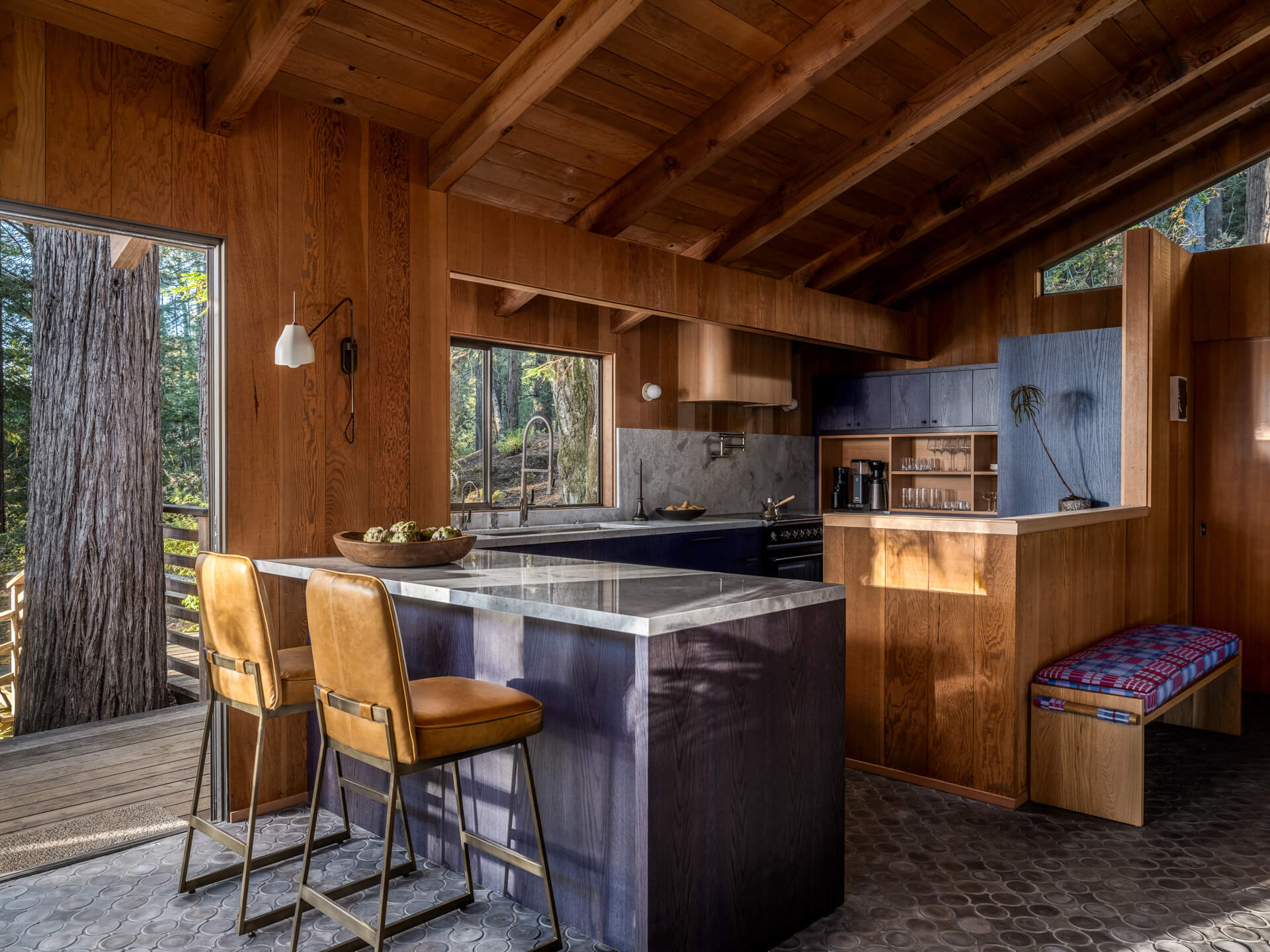 Wood-paneled kitchen with exposed ceiling beams, dark stone island, vintage bar stools, and forest views.
