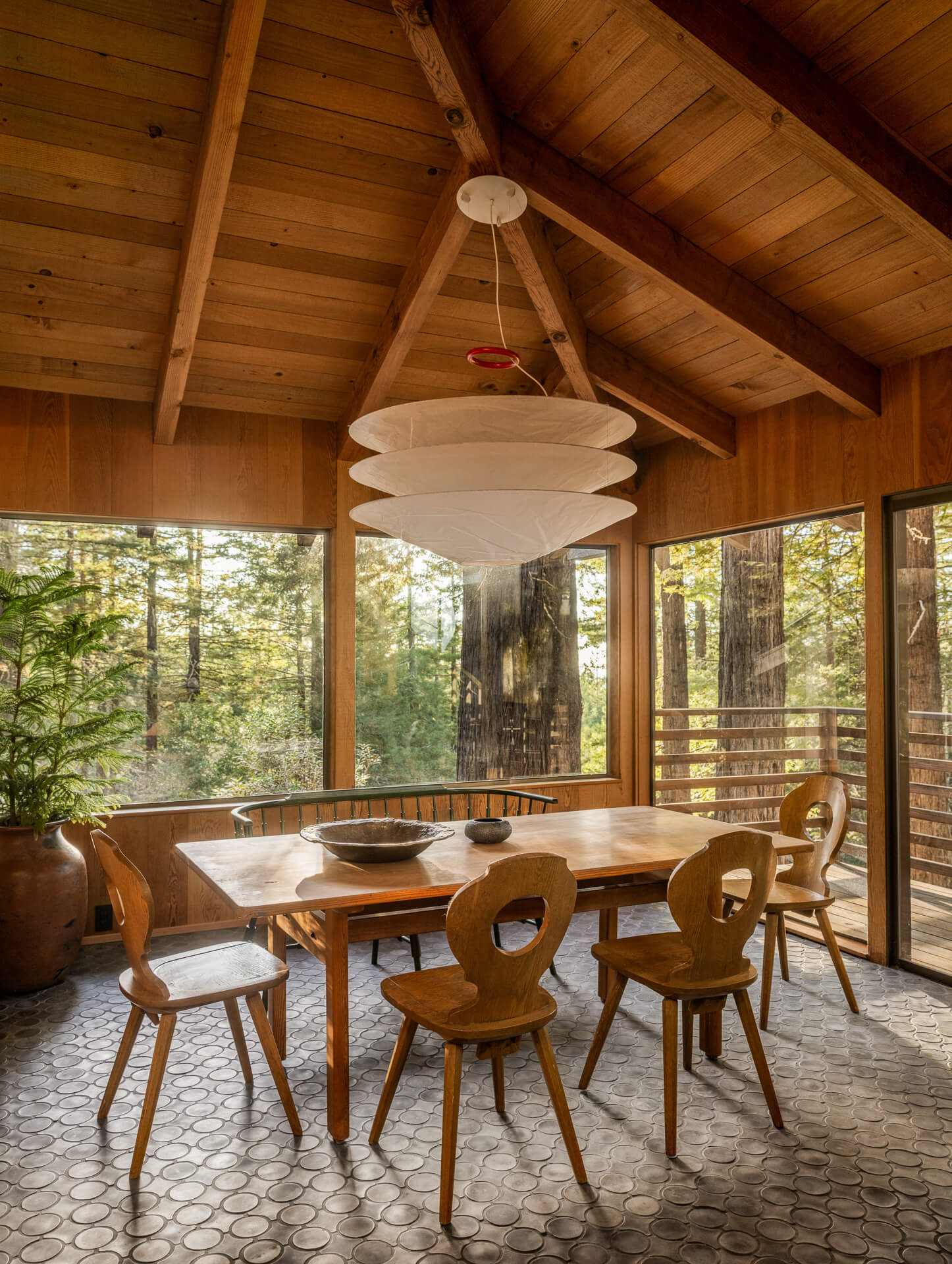 Dining room with wood-paneled ceiling, sculptural dining chairs, pendant light, and floor-to-ceiling windows overlooking the trees.