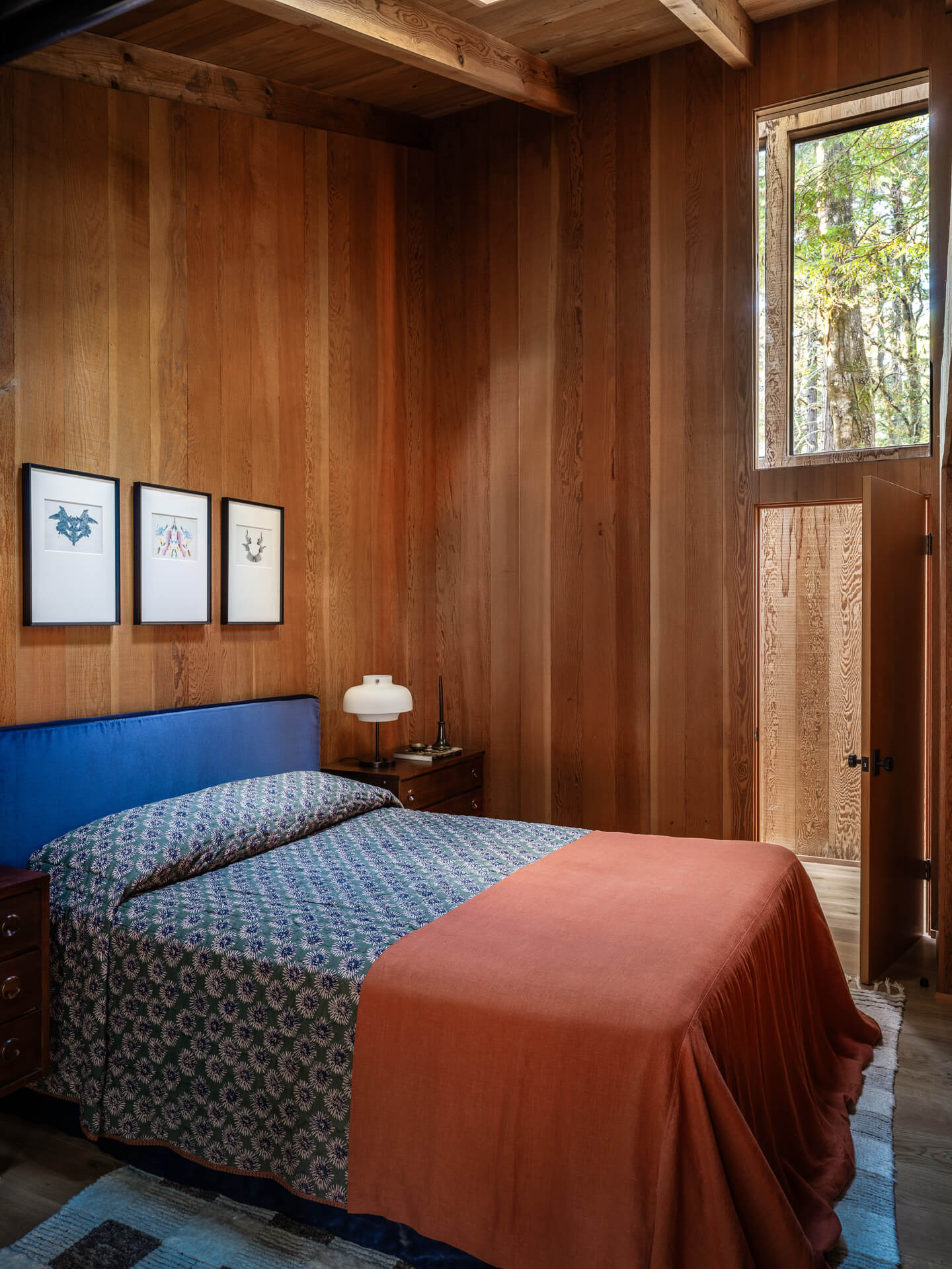 Cozy wood-paneled bedroom with a blue patterned bed, rust-colored throw, framed artwork, and tall window.