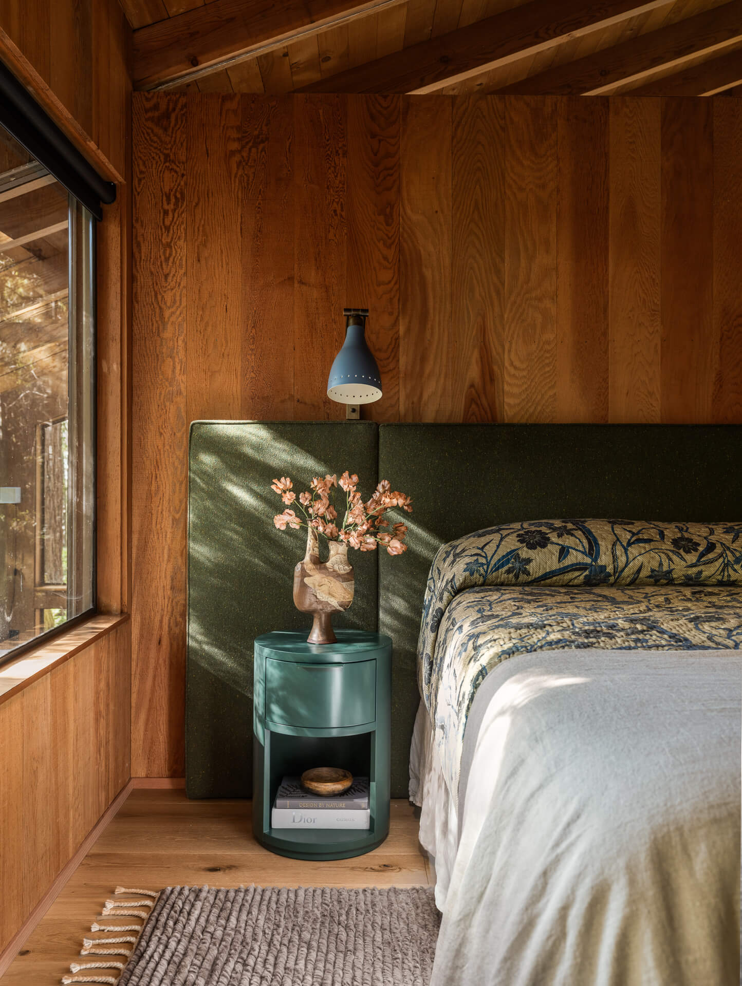 Warm wood-paneled bedroom with green upholstered headboard, patterned bedding, vintage side table, and forest-facing window.