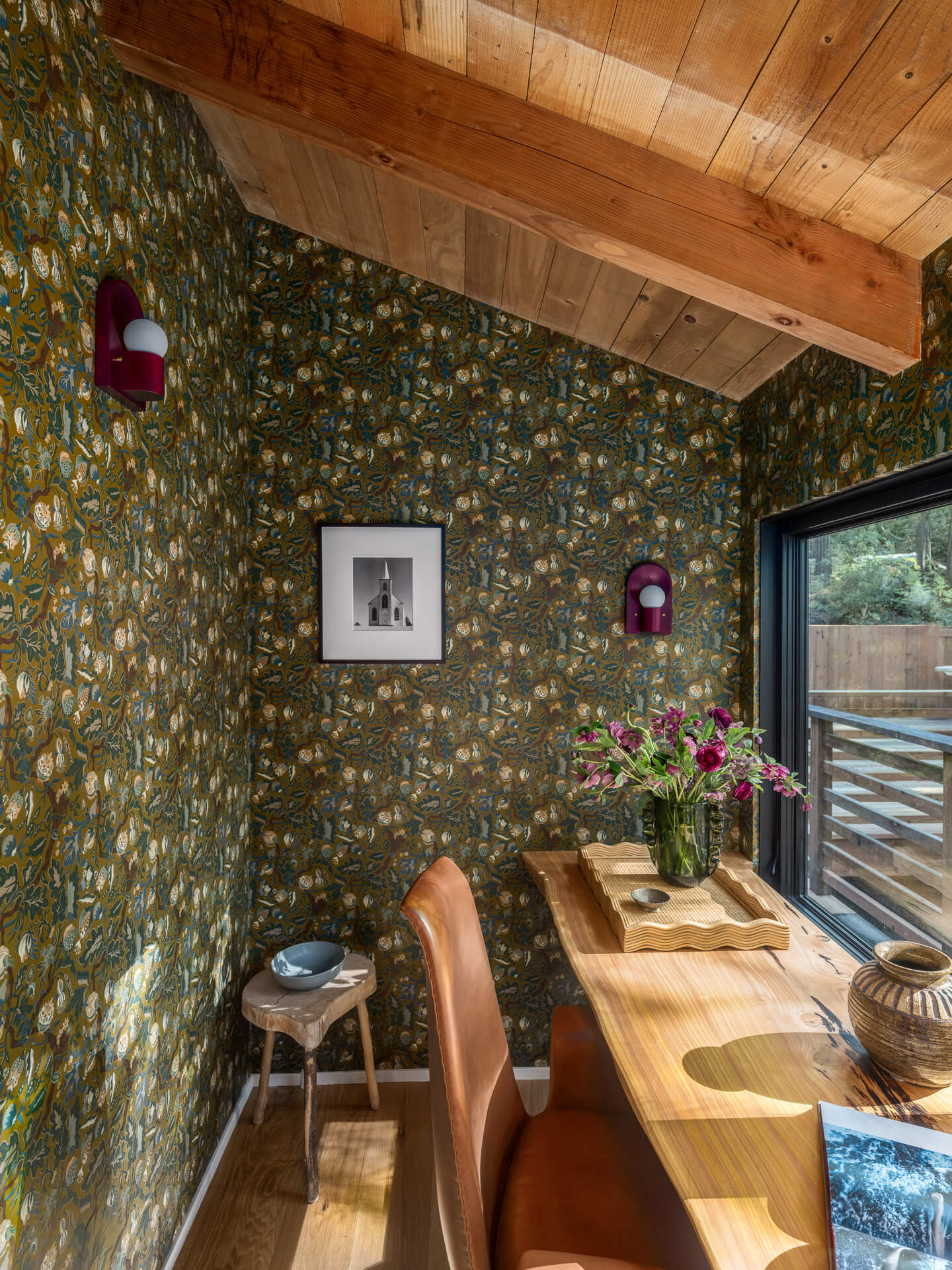 Office nook with floral wallpaper, wood table and banquette seating, colorful pendant lights, and windows to the trees.