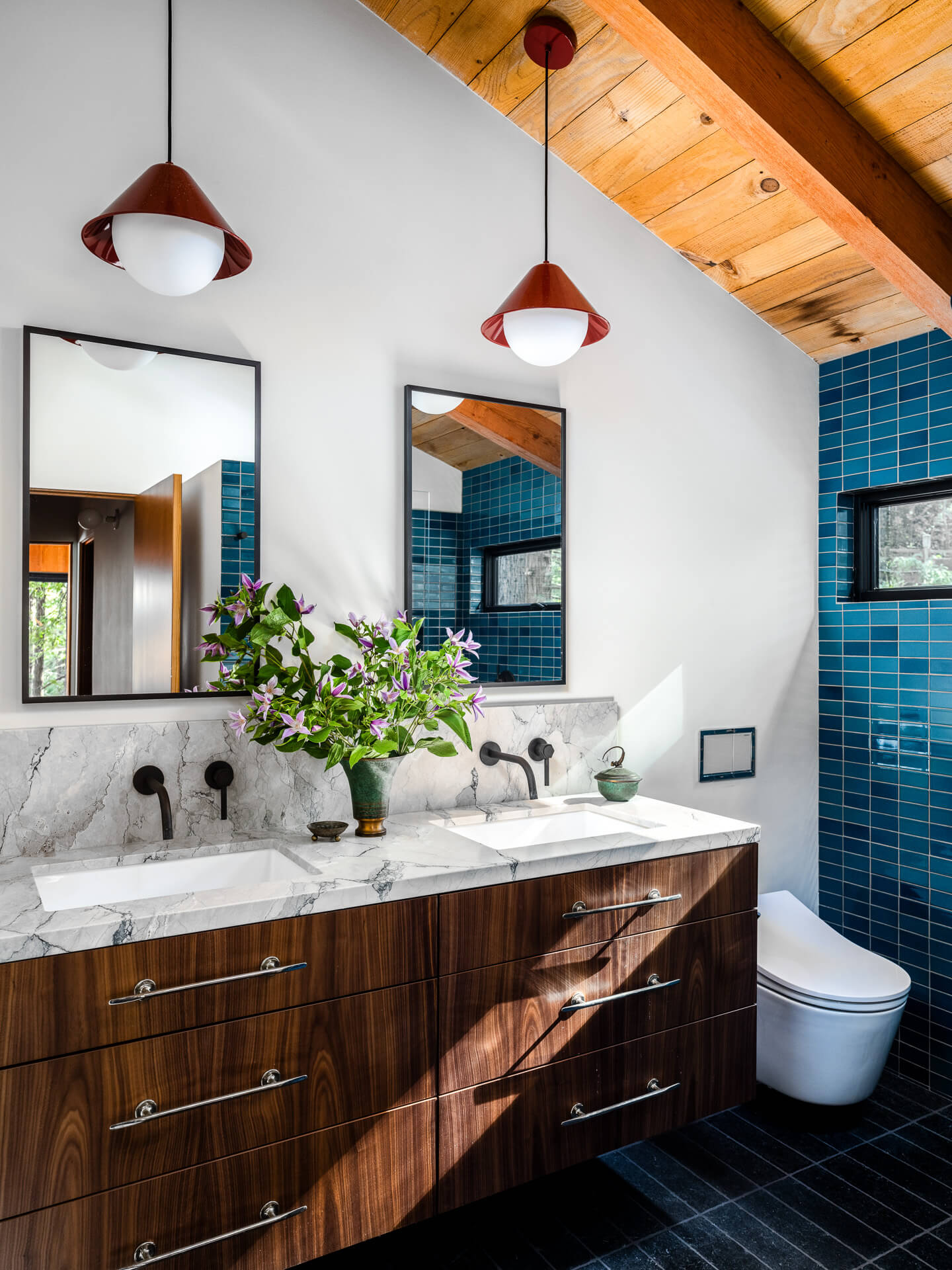 Bright bathroom with walnut vanity, marble countertop, blue tile shower, red pendant lights, and exposed wood ceiling.