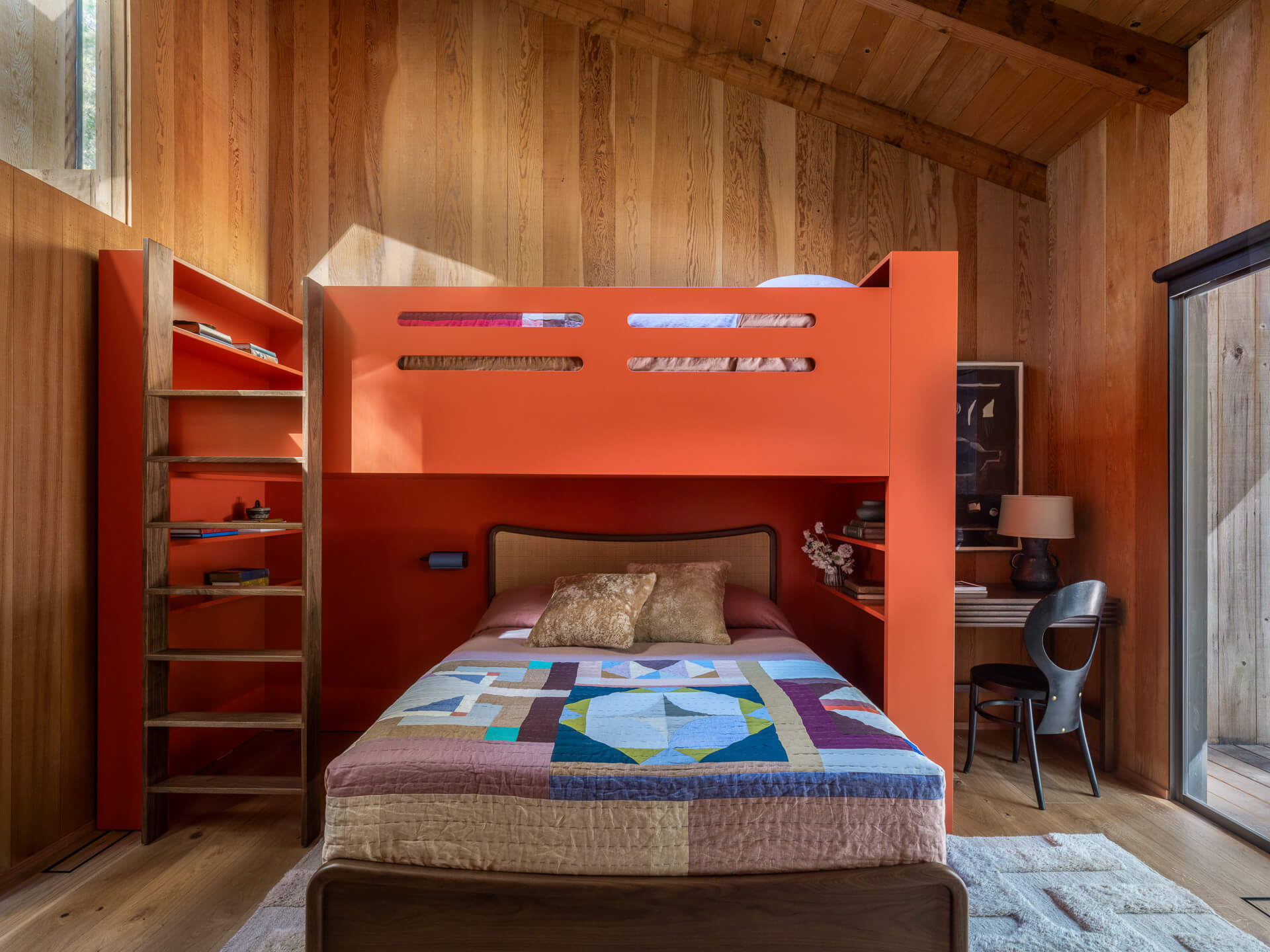Children’s bedroom with bold orange built-in bunk bed, colorful quilt, wood-paneled walls, and exposed ceiling beams.