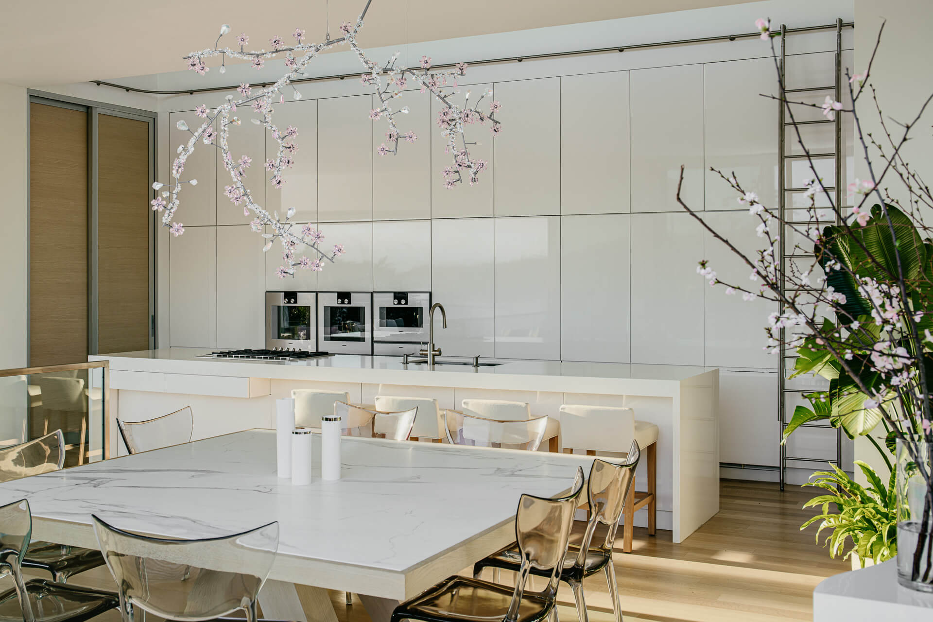 White kitchen and dining area with marble table, transparent chairs, flowering branches, and sleek built-in cabinetry.