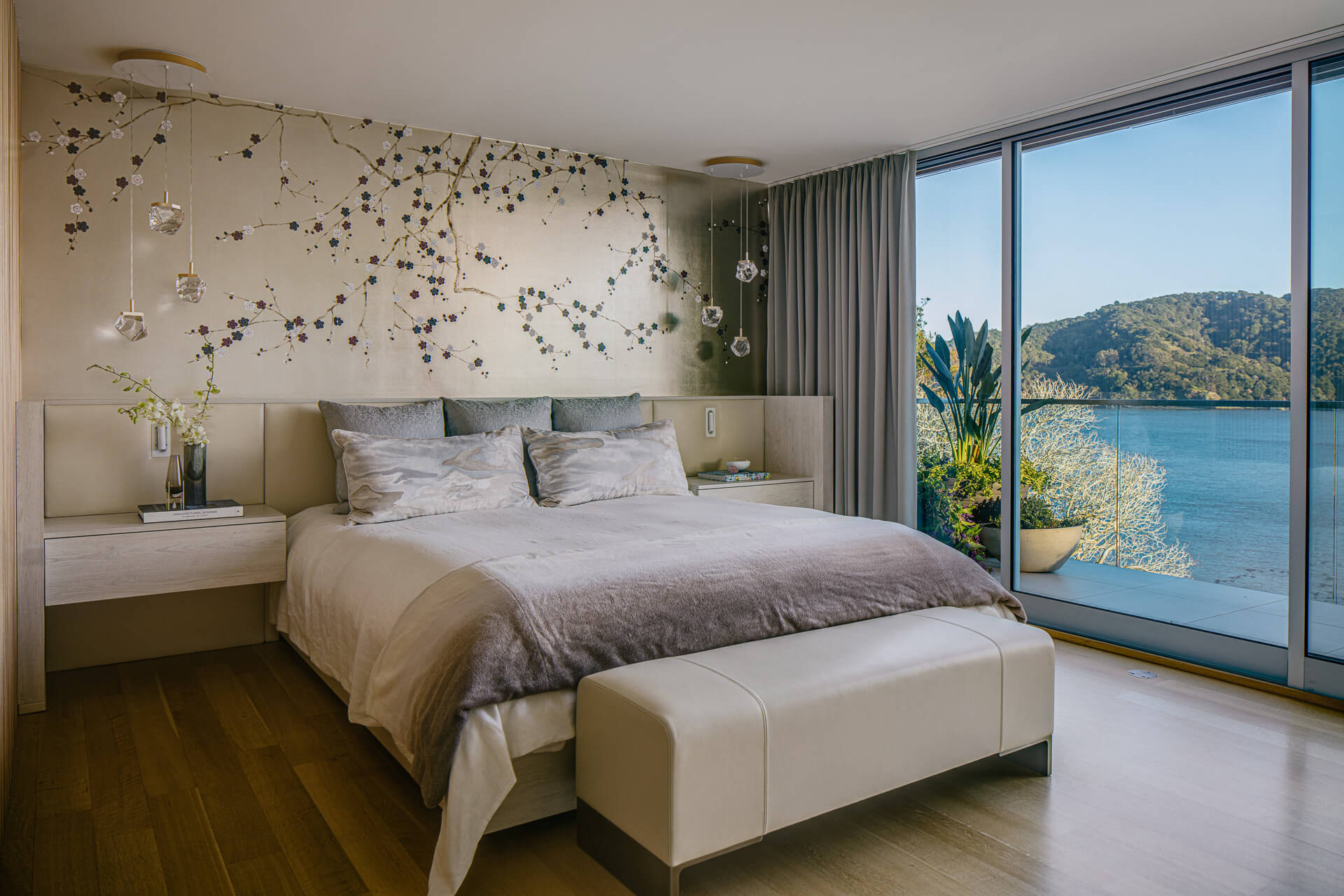 Serene bedroom with upholstered bed, suspended decorative installation, and expansive windows overlooking the water.