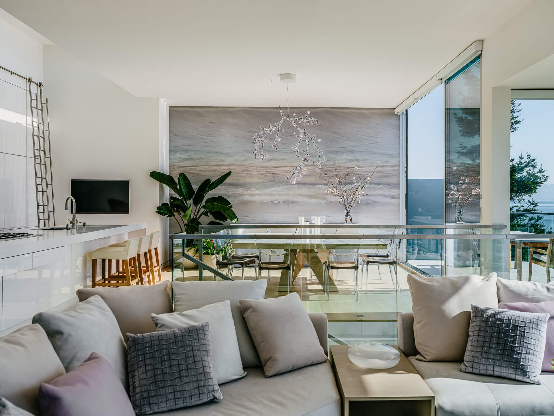Open-plan living and dining space with pale sectional seating, glass railings, and floor-to-ceiling water views.