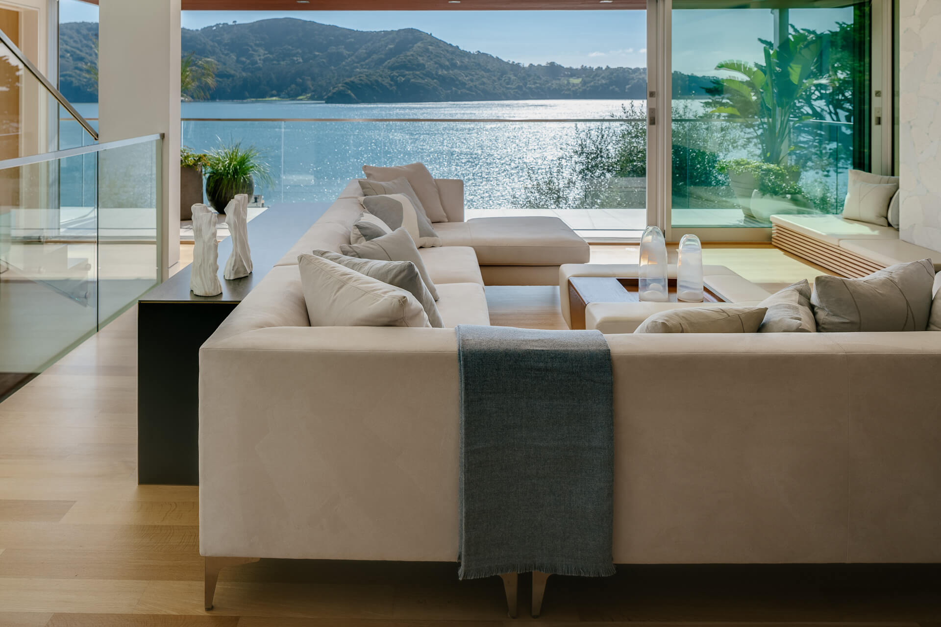 Waterfront living room with large cream sectional, layered tables, and broad windows overlooking the bay and hills.