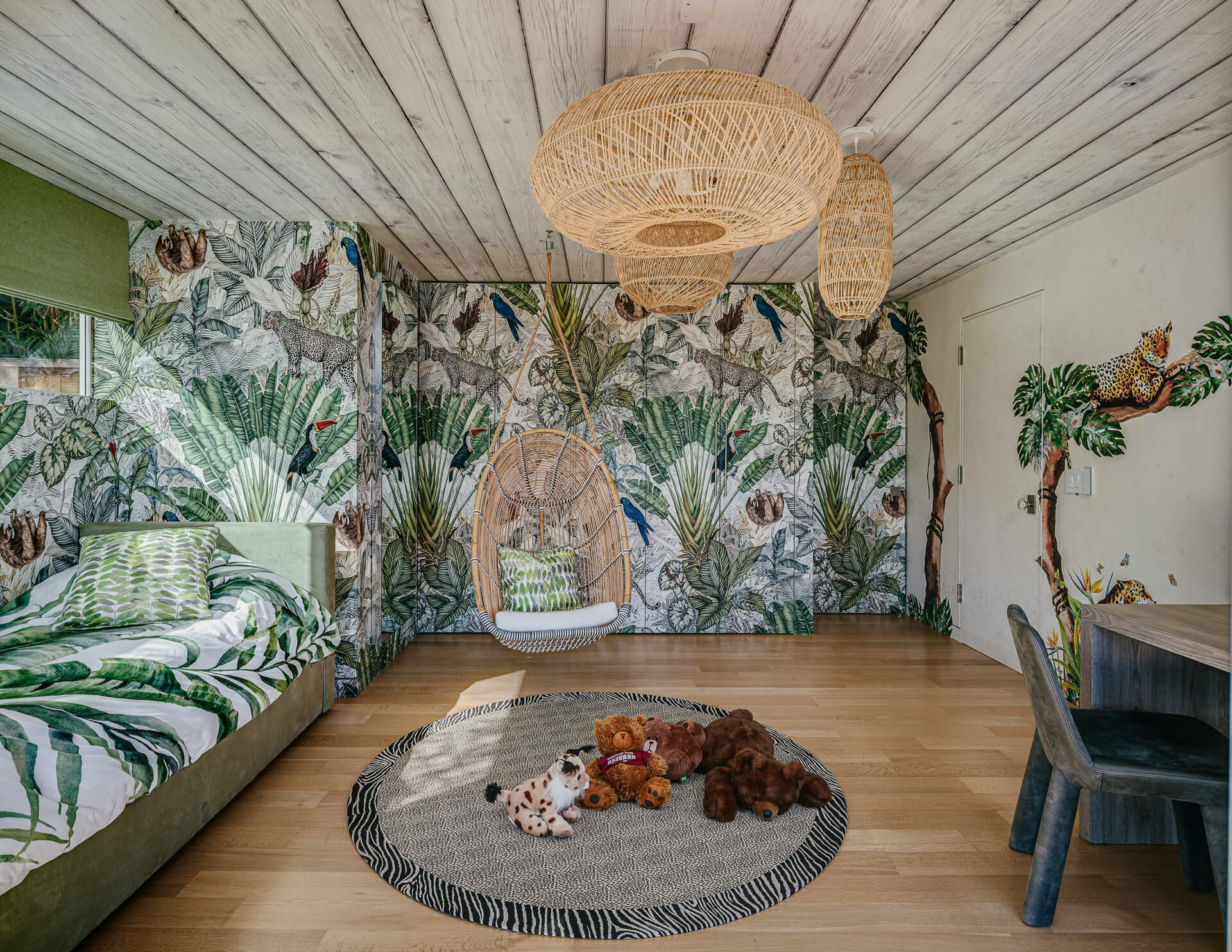 Children’s room with tropical mural walls, hanging wicker chair, daybed, woven lights, and stuffed animals.