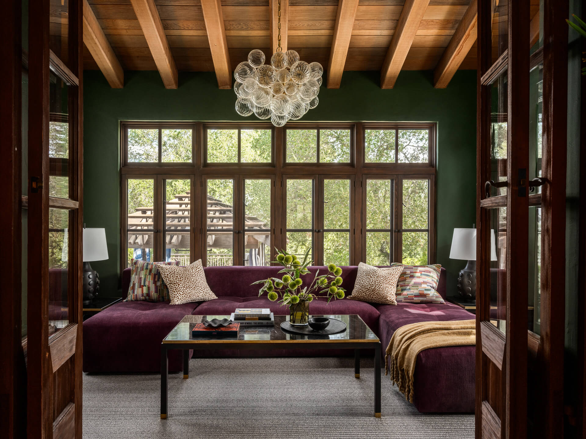 Deep green sitting room with purple sectional sofa, exposed wood ceiling beams, large windows, and sculptural glass chandelier.