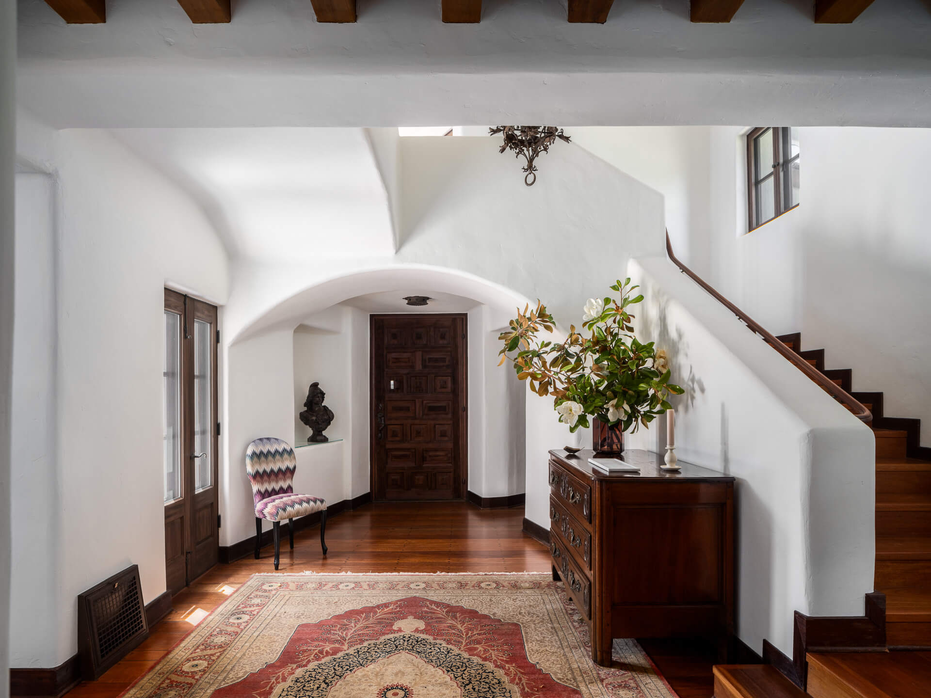 Bright entry hall with white arched walls, wood staircase, patterned rug, and a console topped with flowers.
