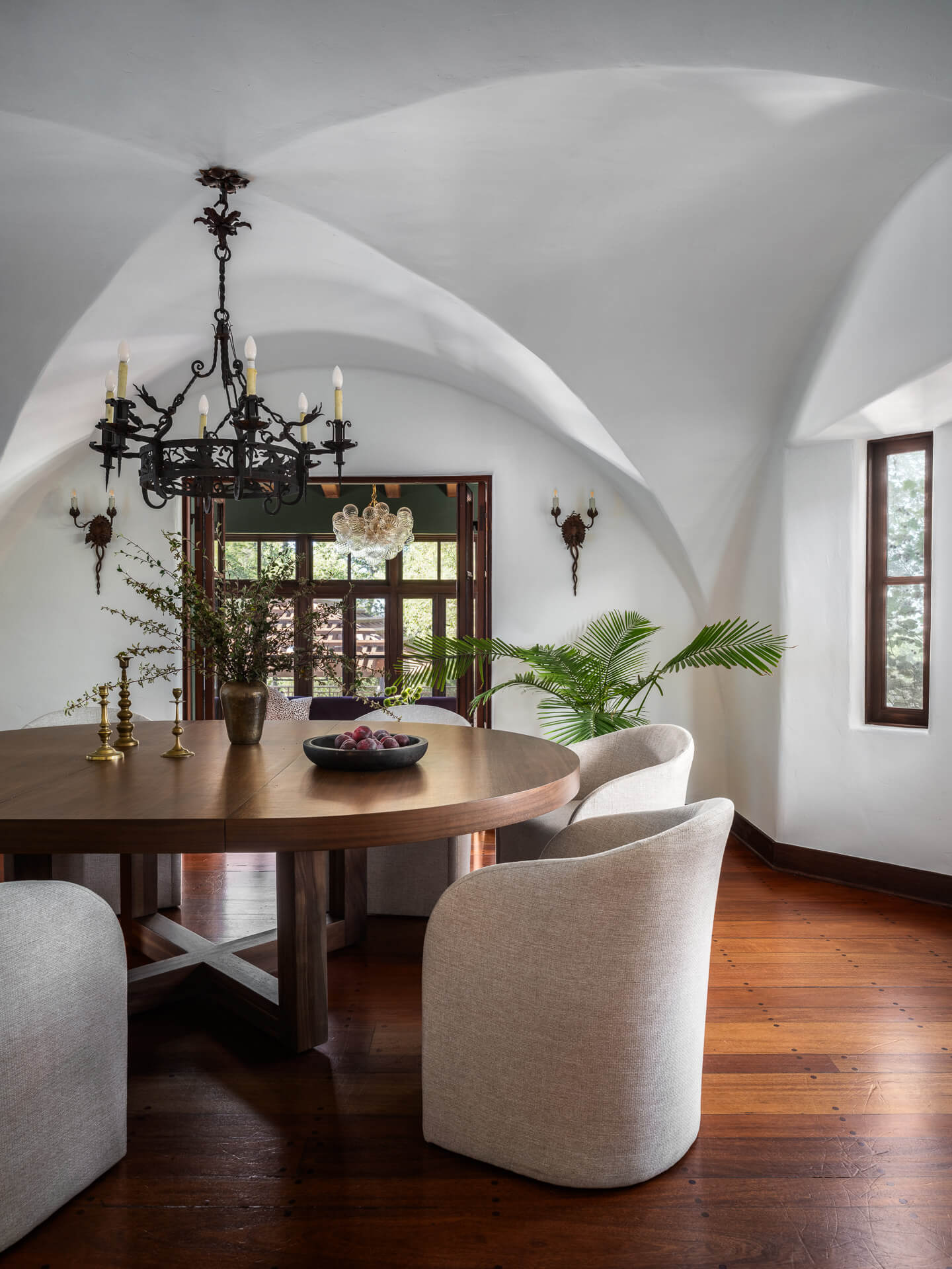 Dining room with round wood table, upholstered chairs, vaulted white ceiling, and ornate black chandelier.