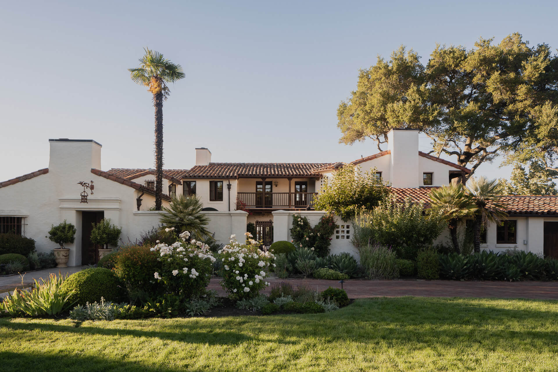 Spanish-style white stucco home with tiled roof surrounded by lawn, mature trees, and garden plantings.
