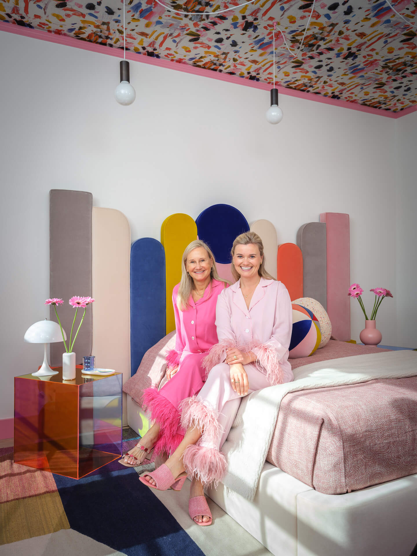 Two women seated together in a colorful bedroom with a sculptural multicolored headboard.