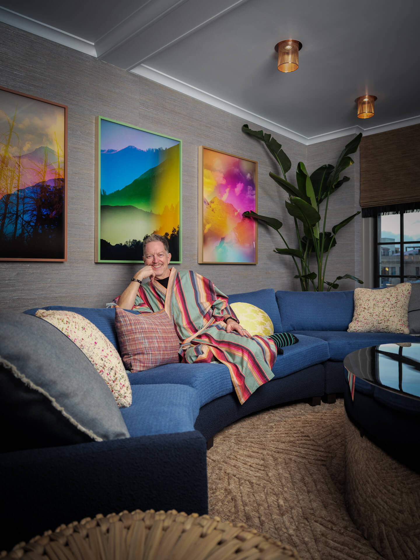 John Anderson seated on a blue sofa beneath a trio of colorful artworks.