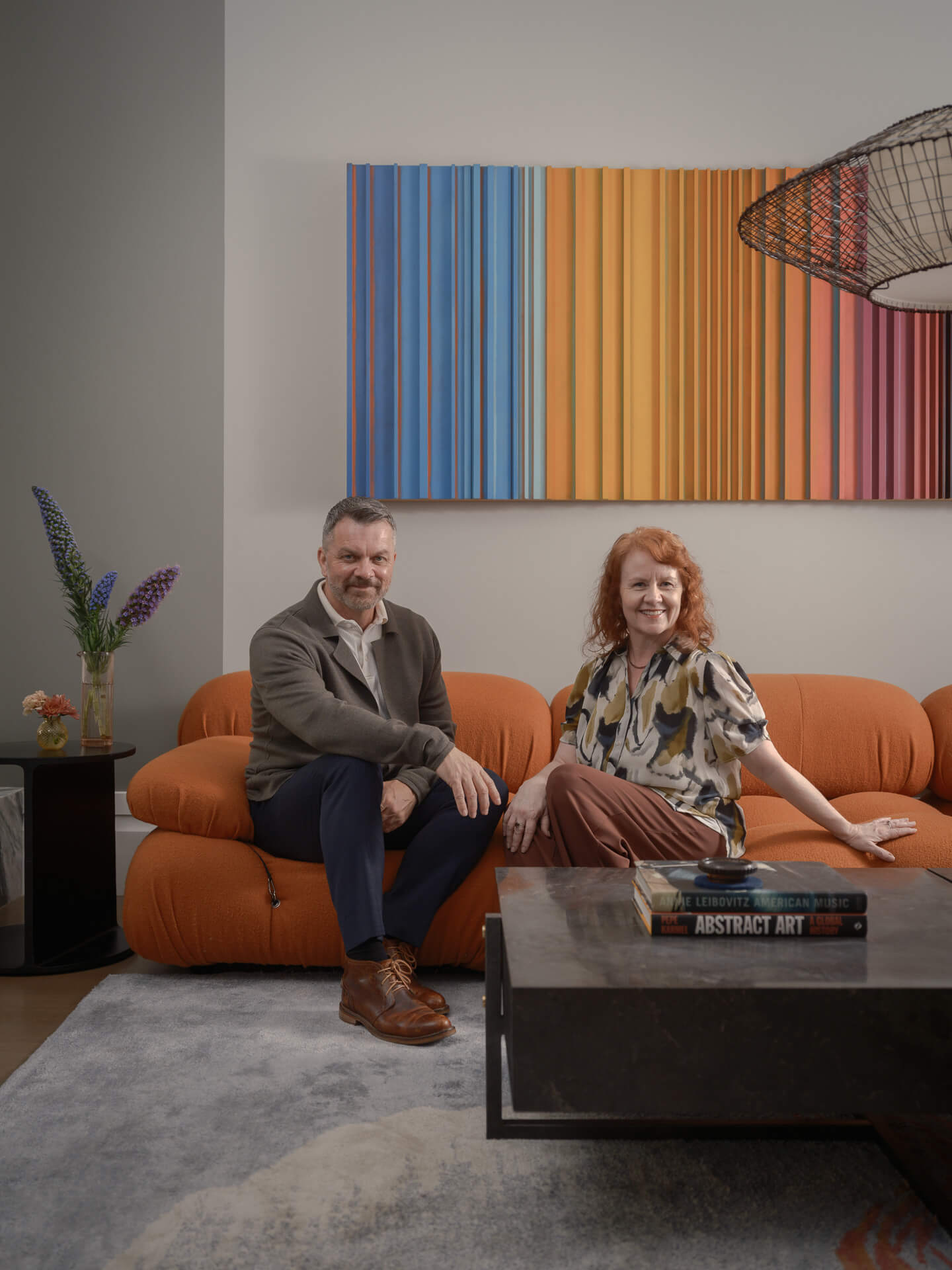 Two LOCZI Design principals seated on an orange sofa beneath colorful striped artwork.