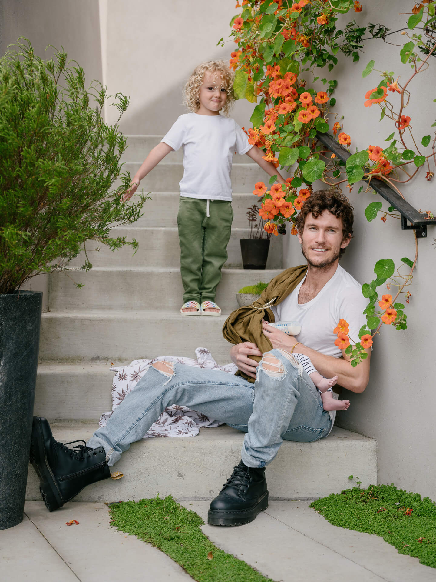 Sean Leffers seated on outdoor steps with a child, surrounded by greenery and orange flowers.