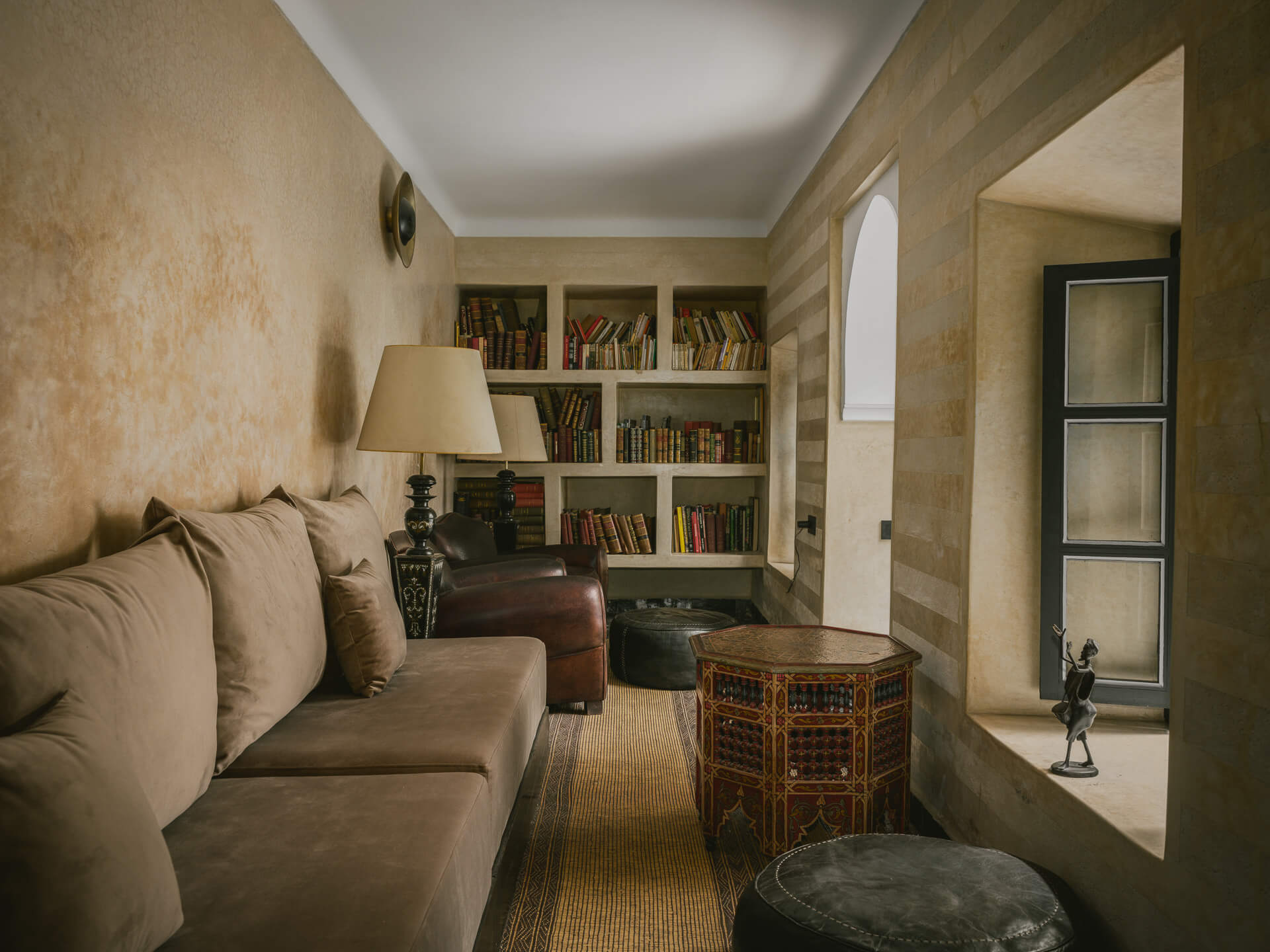 Narrow sitting room with built-in sofa, bookshelves, woven table, and a deep window opening.
