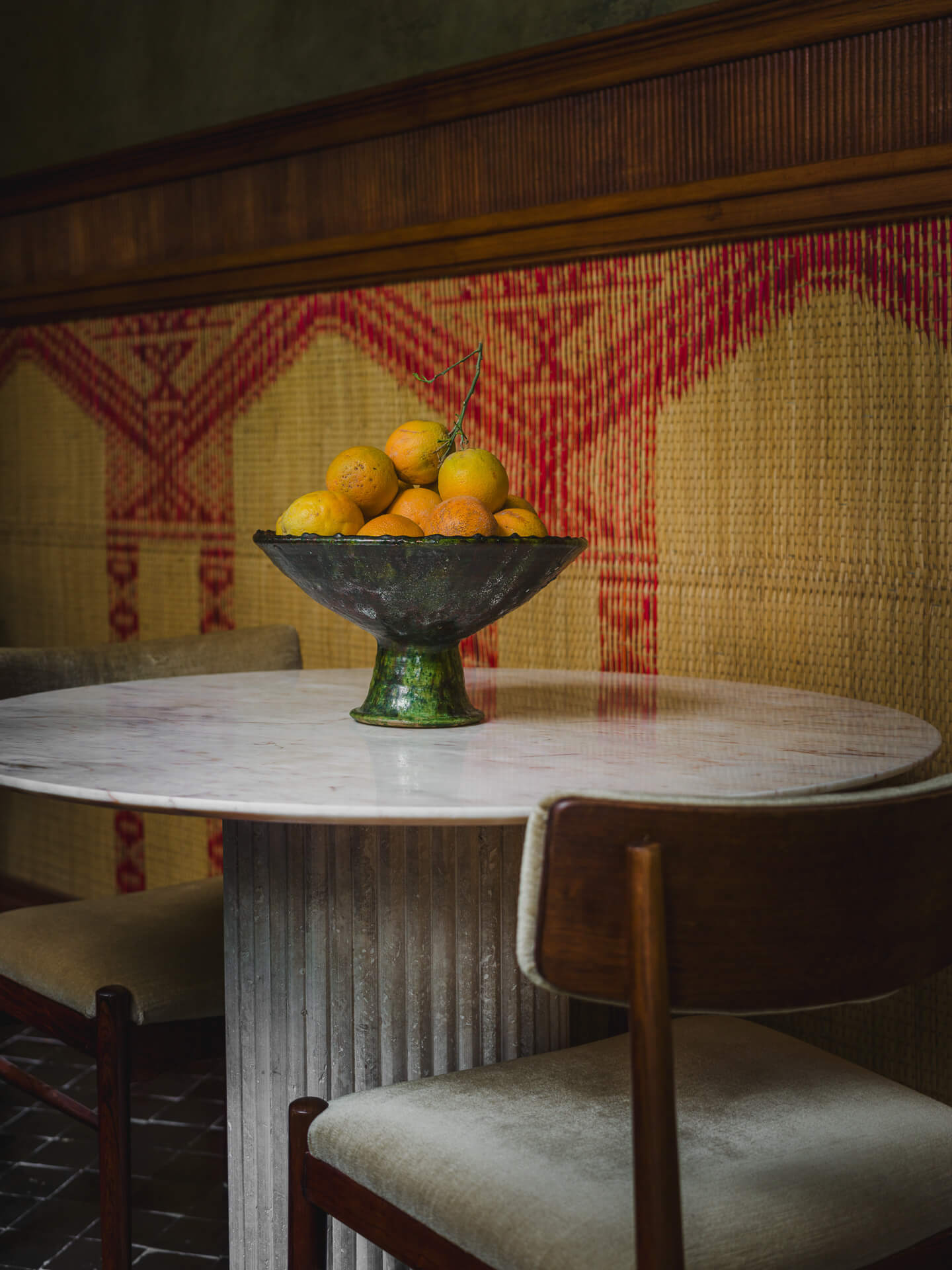 Round stone dining table with a bowl of citrus against patterned woven wall panels.