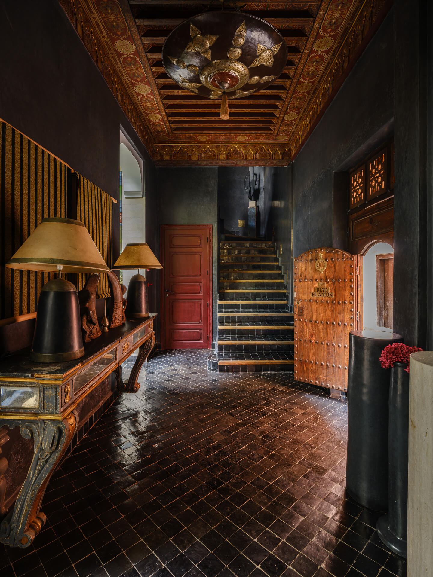 Dark entry hall with carved wood details, console tables, lamps, and a staircase beyond.