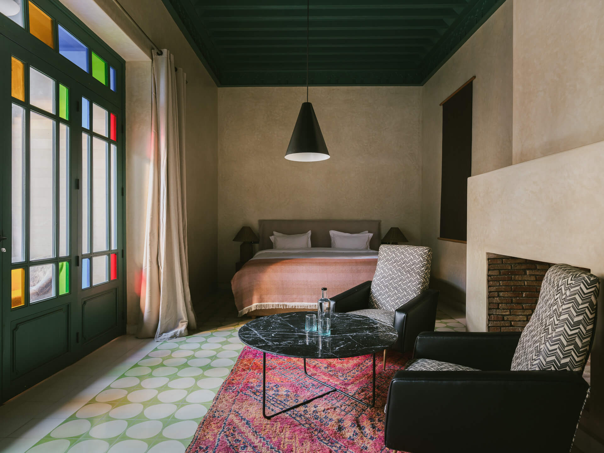 Bedroom sitting area with green ceiling, colorful glass doors, patterned rug, and dark lounge chairs.