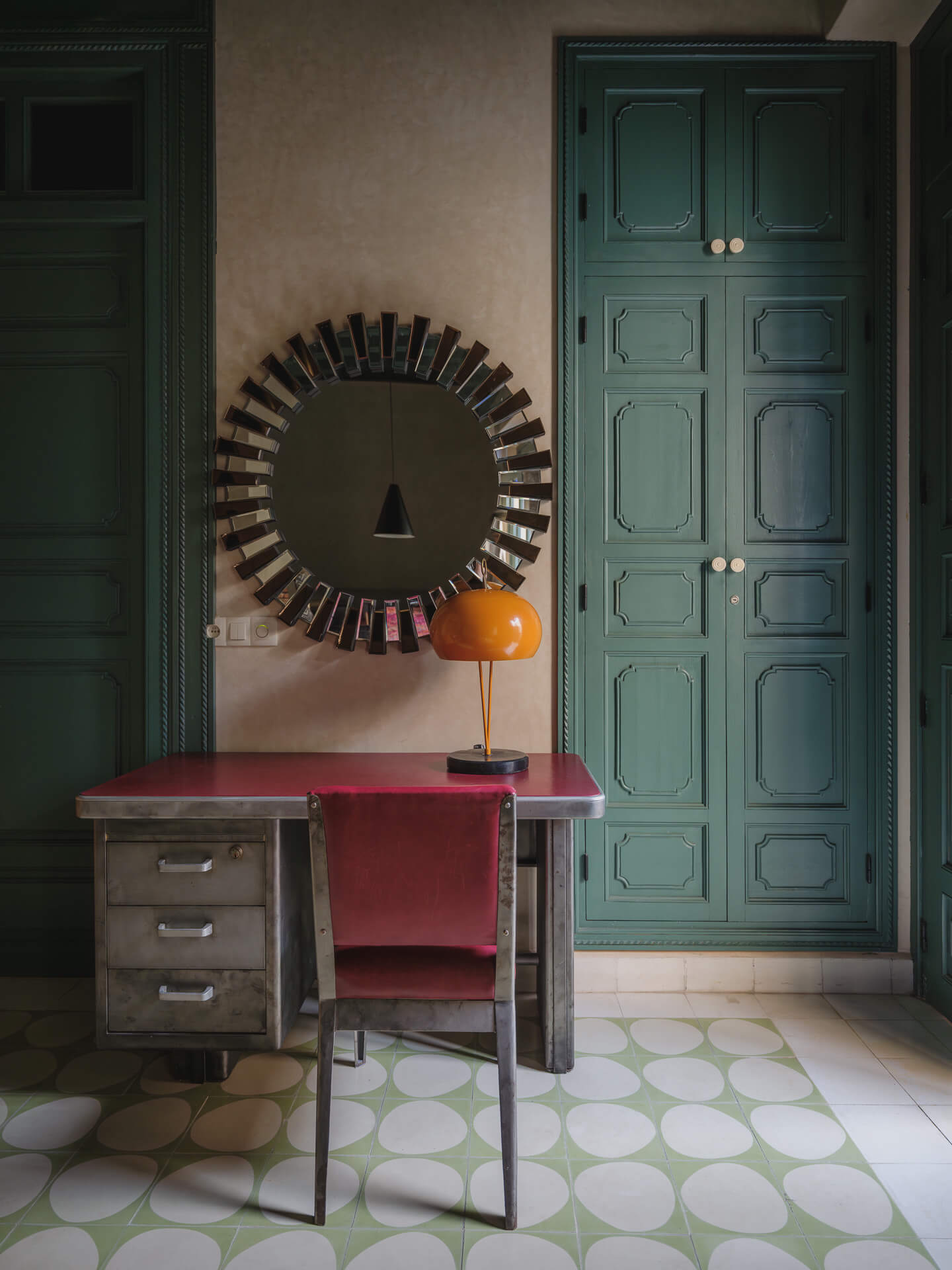 Small desk beneath a round decorative mirror beside tall teal-painted doors.